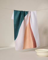 The GEOMETRY Fan kitchen towel features abstract wavy patterns in dark green, peach, and rust. Made from recycled materials for eco-friendly style, it hangs above a white ceramic dish and bowl on a neutral background.