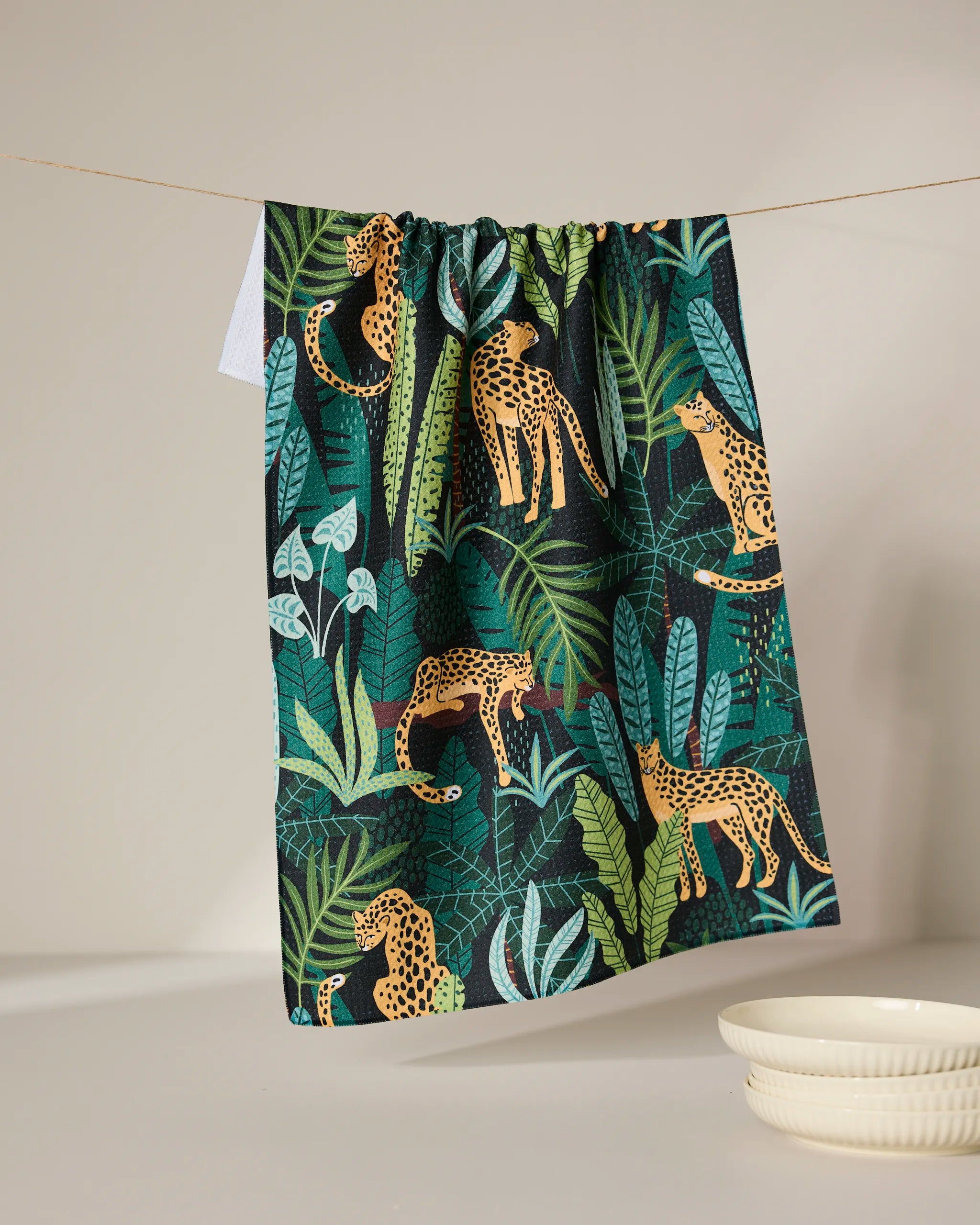 The GEOMETRY Feeling Spotty tea towel, featuring a vibrant jungle pattern with cheetahs and lush greenery, hangs on a line above a cream bowl against a neutral backdrop.