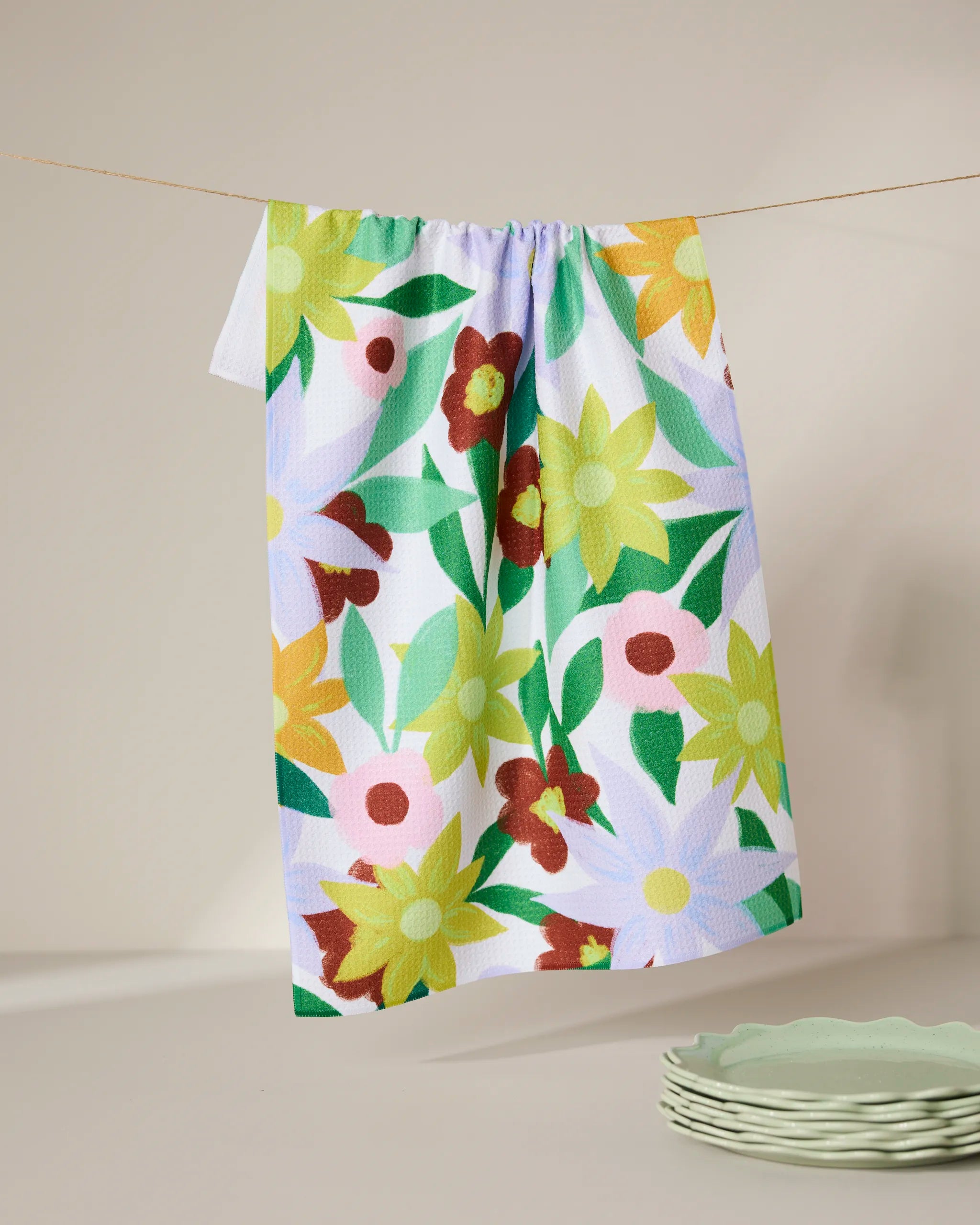 The GEOMETRY Floral Fields tea towel, featuring a vibrant yellow, green, red, and white floral design, hangs on a clothesline above pale green plates set against a neutral background.