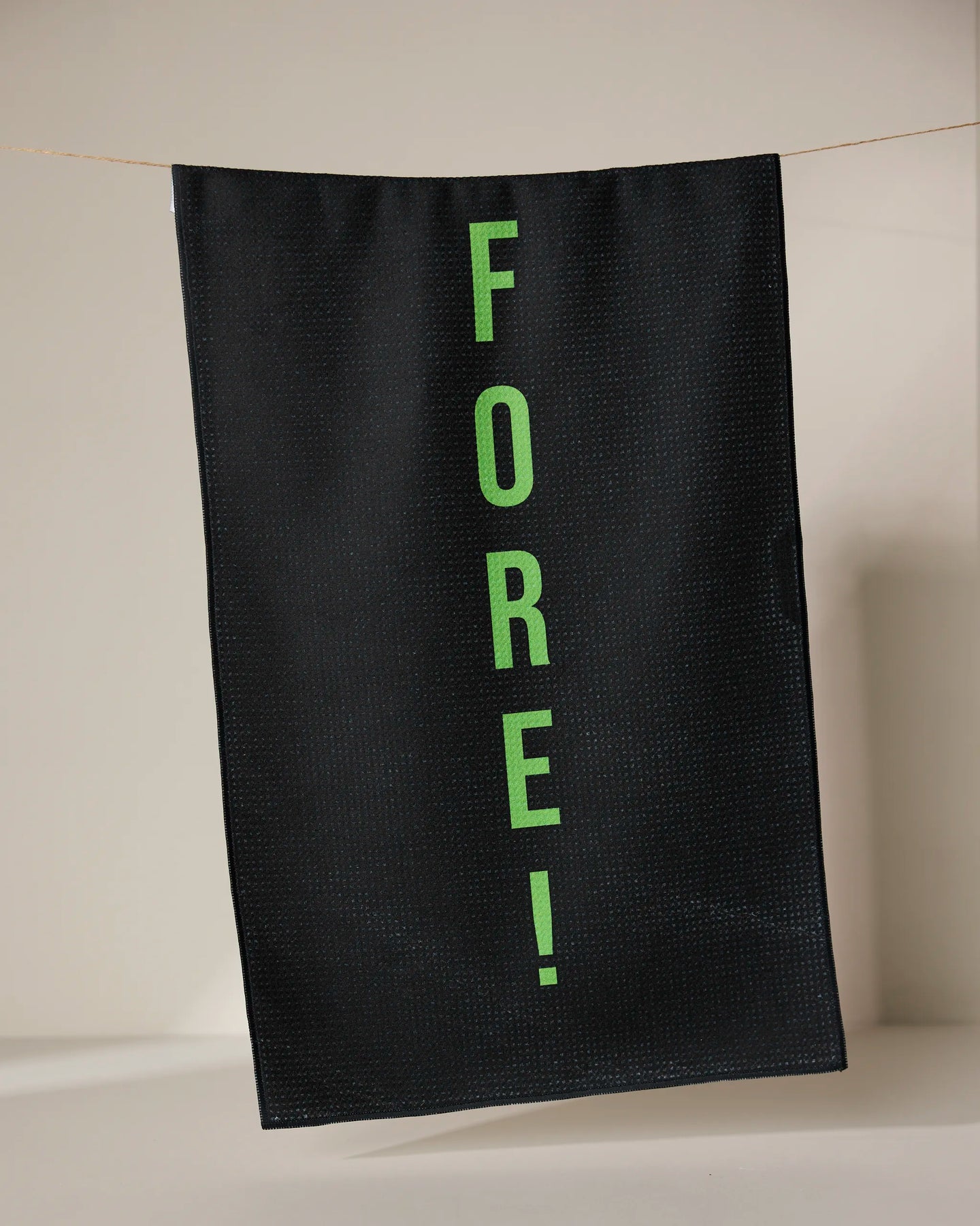 The GEOMETRY Fore tea towel, made from recycled materials, dries quickly and features FORE! in bright green letters printed vertically on black fabric, hanging from a thin line against a neutral background.