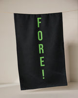 The GEOMETRY Fore tea towel, made from recycled materials, dries quickly and features FORE! in bright green letters printed vertically on black fabric, hanging from a thin line against a neutral background.