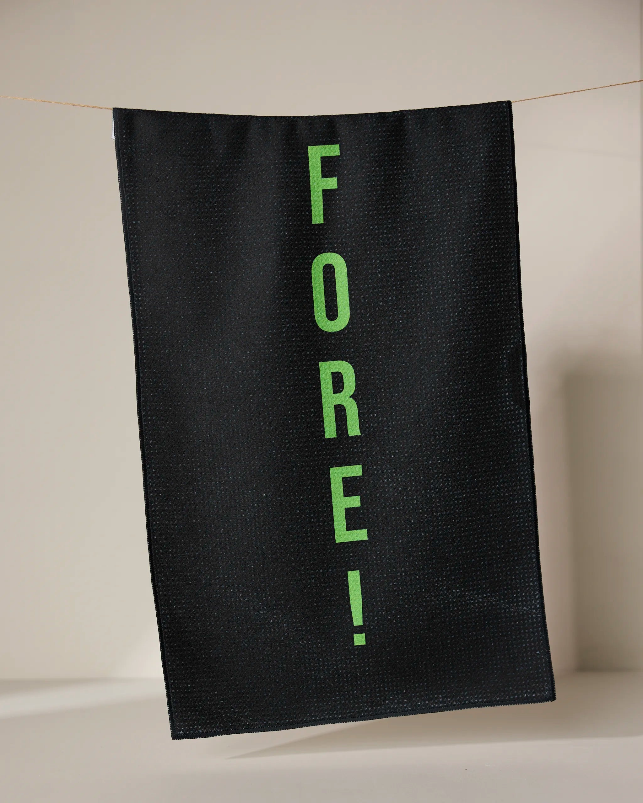 The GEOMETRY Fore tea towel, made from recycled materials, dries quickly and features FORE! in bright green letters printed vertically on black fabric, hanging from a thin line against a neutral background.