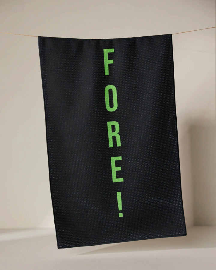 The GEOMETRY Fore tea towel, made from recycled materials, dries quickly and features FORE! in bright green letters printed vertically on black fabric, hanging from a thin line against a neutral background.