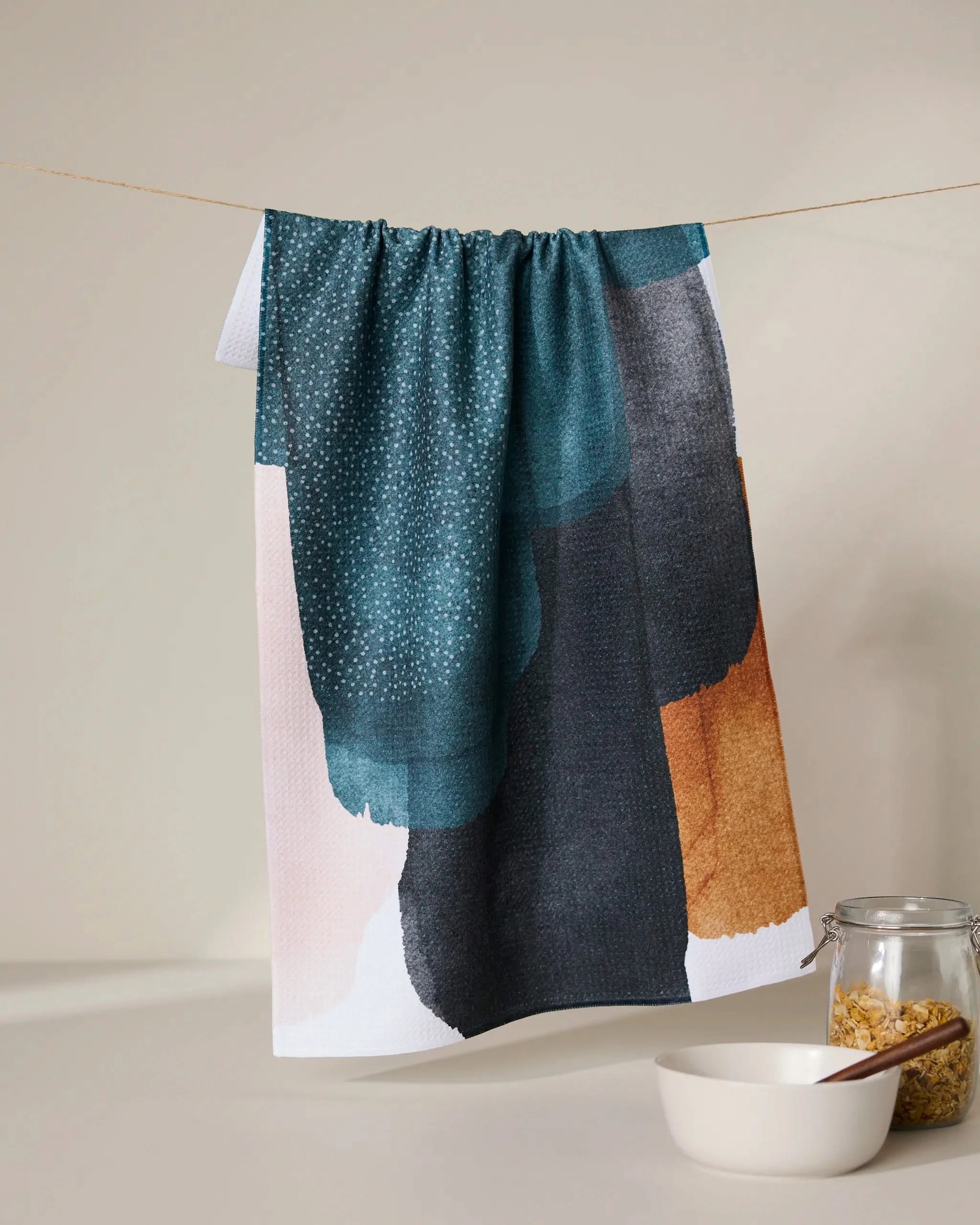 The GEOMETRY Founded tea towel, featuring abstract blue, black, white, and brown brushstrokes, hangs on a string above a white bowl and a glass jar of cereal on a beige surface with a neutral background.