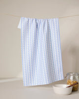 A GEOMETRY Fresh Linen tea towel in light blue and white hangs on a string. Below, a white bowl with wooden spoon, a glass jar of granola, and part of the towel draped to the side complete the scene.