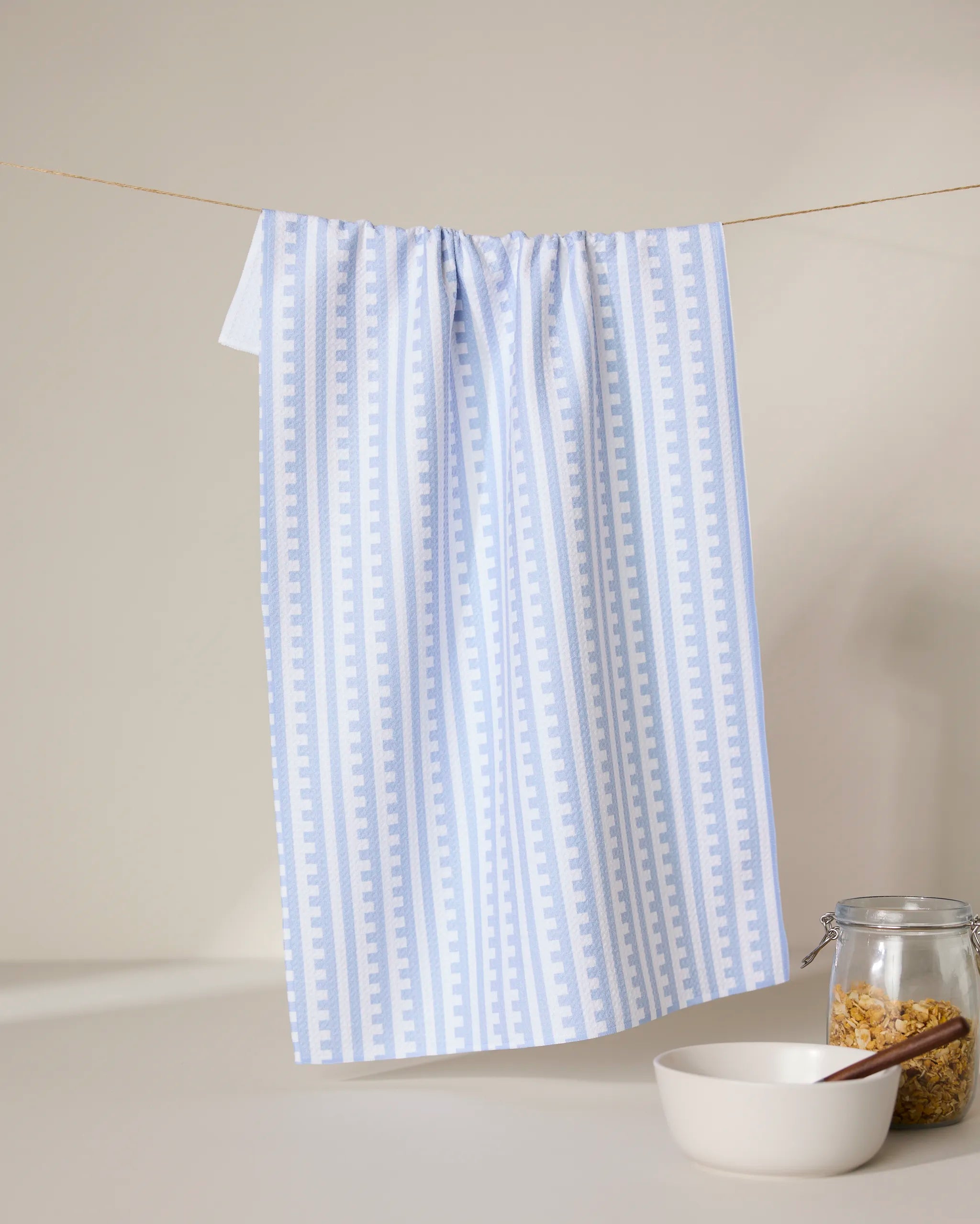 A GEOMETRY Fresh Linen tea towel in light blue and white hangs on a string. Below, a white bowl with wooden spoon, a glass jar of granola, and part of the towel draped to the side complete the scene.