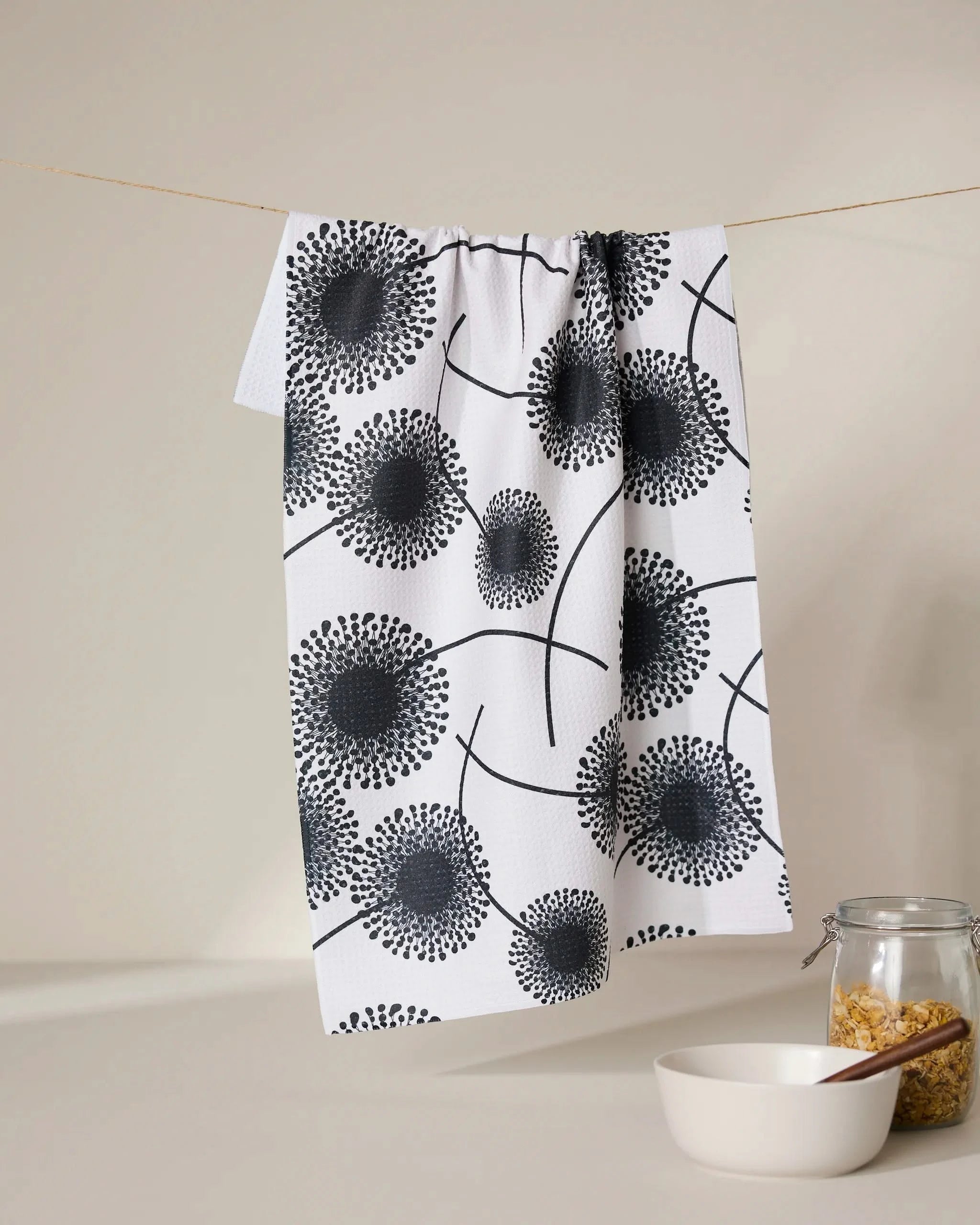 The GEOMETRY Fully Bloomed quick-drying absorbent towel in a black dandelion pattern hangs above a white bowl and cereal-filled jar, beautifully displayed on a light surface with a neutral beige background.