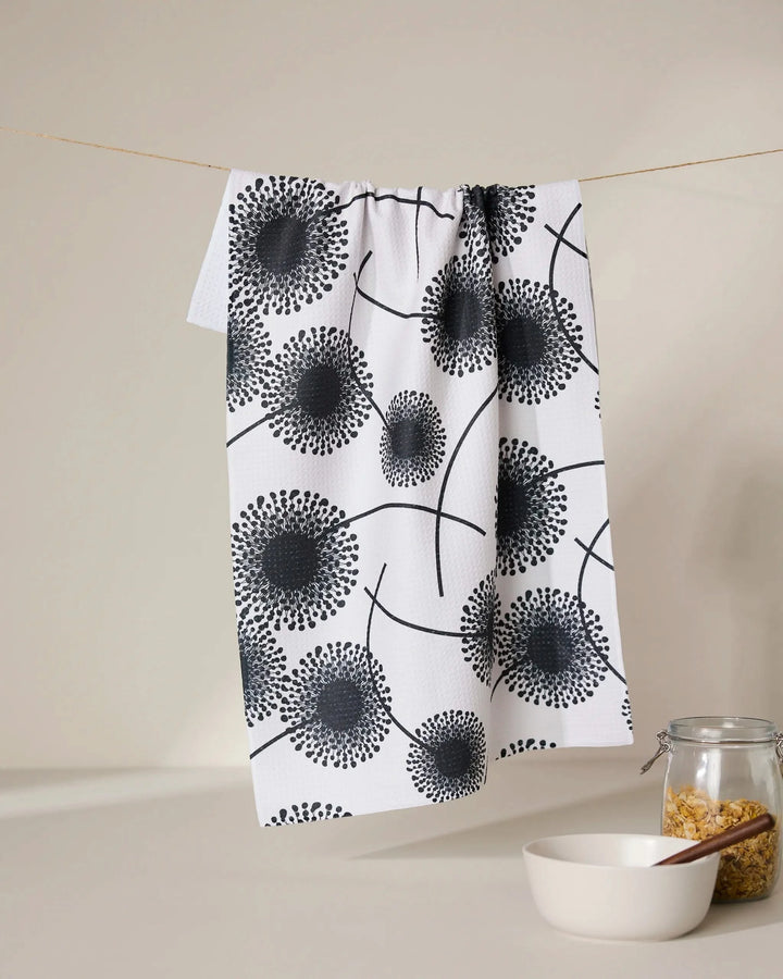 The GEOMETRY Fully Bloomed quick-drying absorbent towel in a black dandelion pattern hangs above a white bowl and cereal-filled jar, beautifully displayed on a light surface with a neutral beige background.