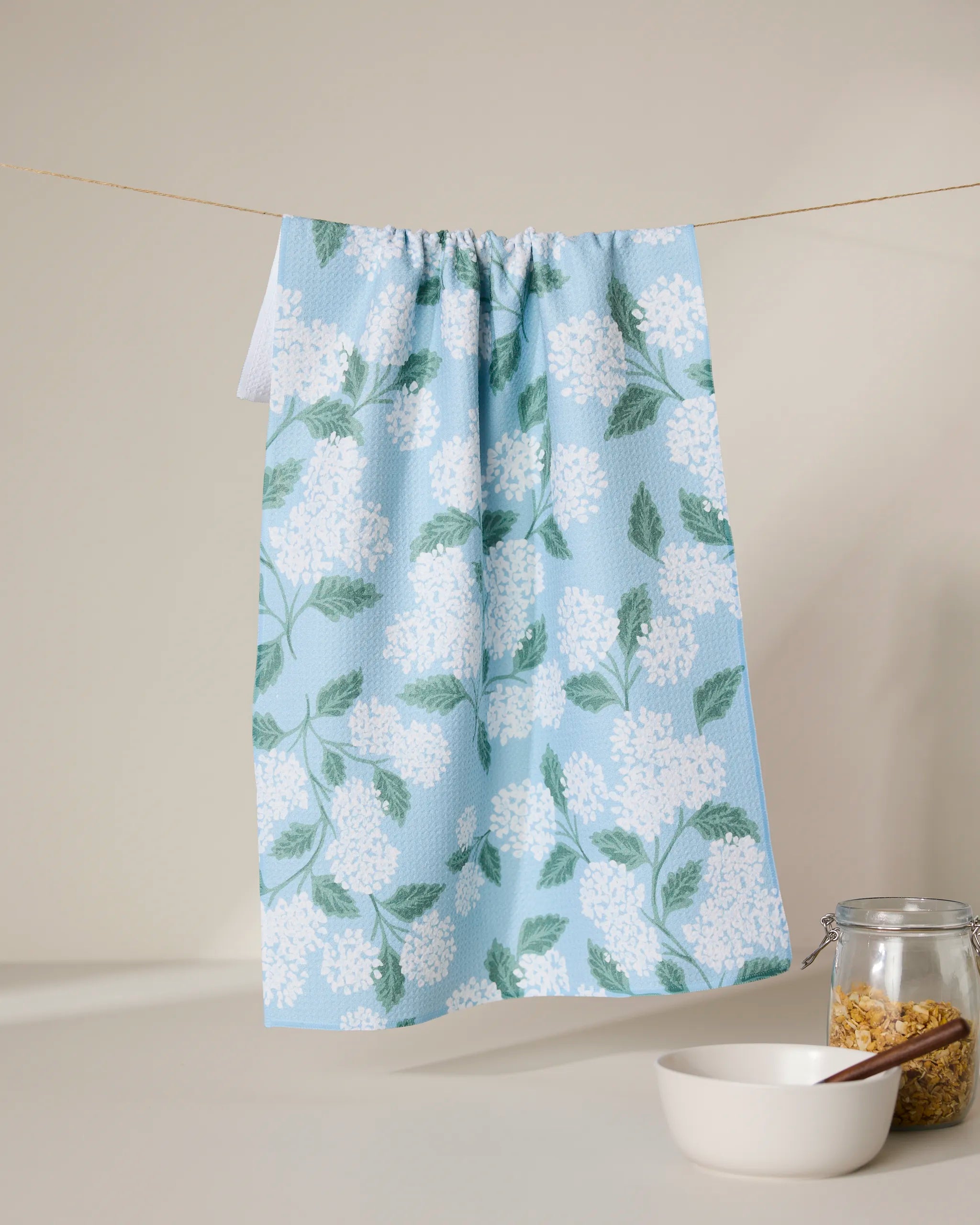 The GEOMETRY Garden Of Edith blue tea towel, featuring a white floral and green leaf pattern, hangs on a string above a white bowl and pasta-filled jar. Made from quick-drying, recycled materials.
