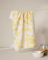 The GEOMETRY Garden of Gold tea towel with a yellow botanical print hangs on a string. Below, a white bowl and a glass jar filled with granola sit on a light surface against a neutral background.