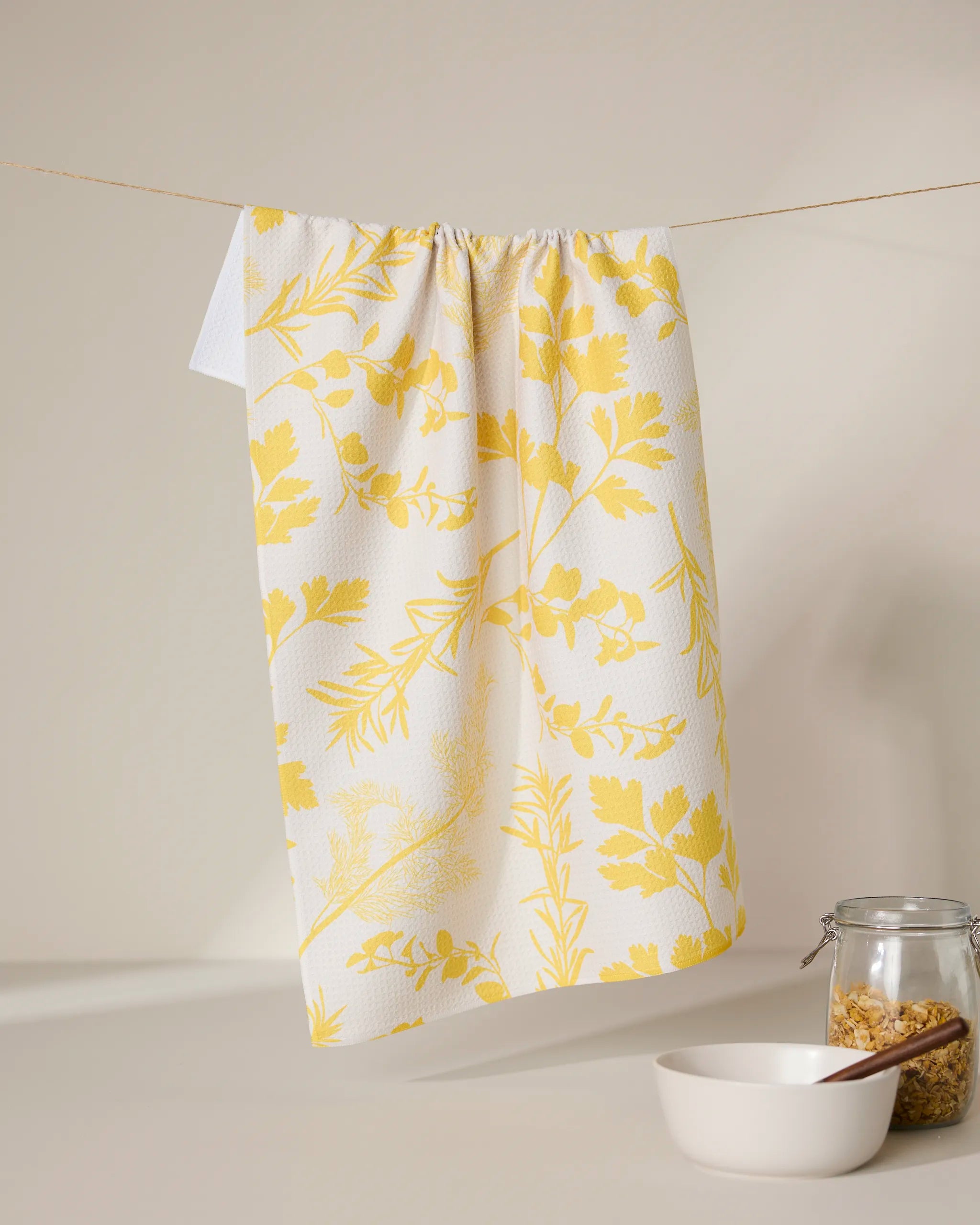 The GEOMETRY Garden of Gold tea towel with a yellow botanical print hangs on a string. Below, a white bowl and a glass jar filled with granola sit on a light surface against a neutral background.