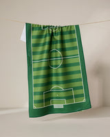 The GEOMETRY Goal tea towel features a green soccer field design with white markings and stripes, hanging from a string against a neutral backdrop.