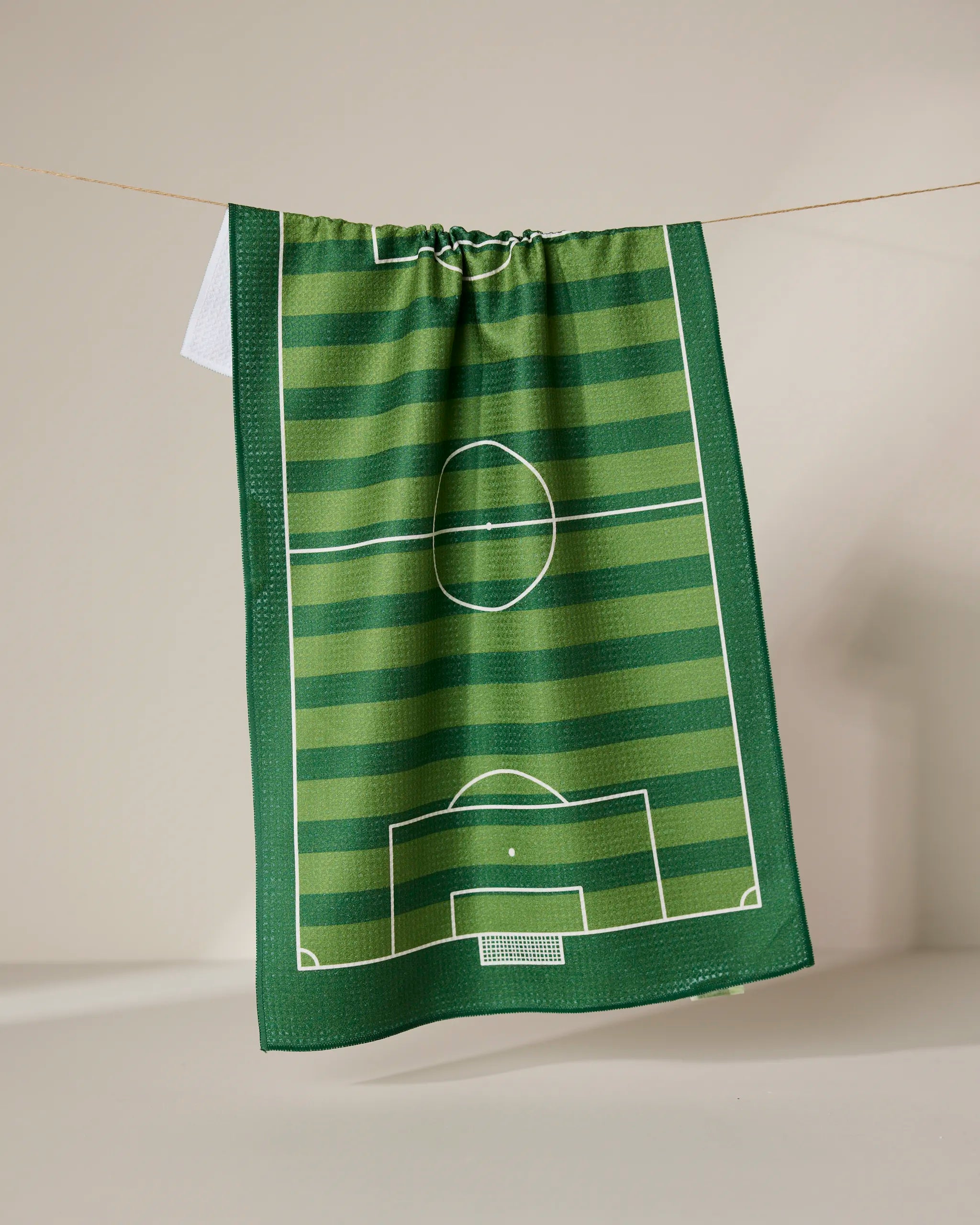 The GEOMETRY Goal tea towel features a green soccer field design with white markings and stripes, hanging from a string against a neutral backdrop.
