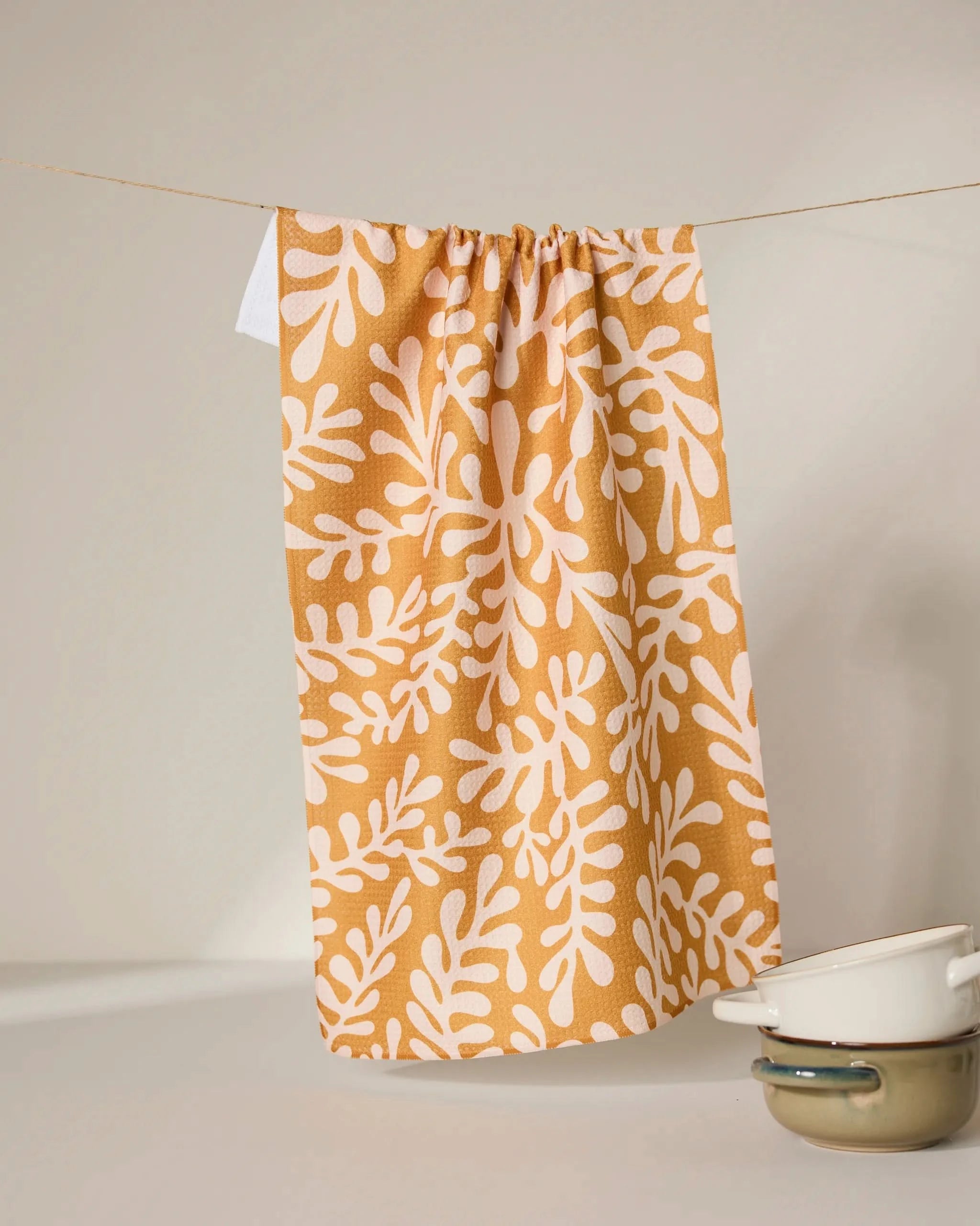 The GEOMETRY Golden Fall tea towel, crafted from recycled materials in mustard yellow with a white leafy pattern, hangs on a string. In front, two cream enamel pots with dark rims are stacked against a neutral background.