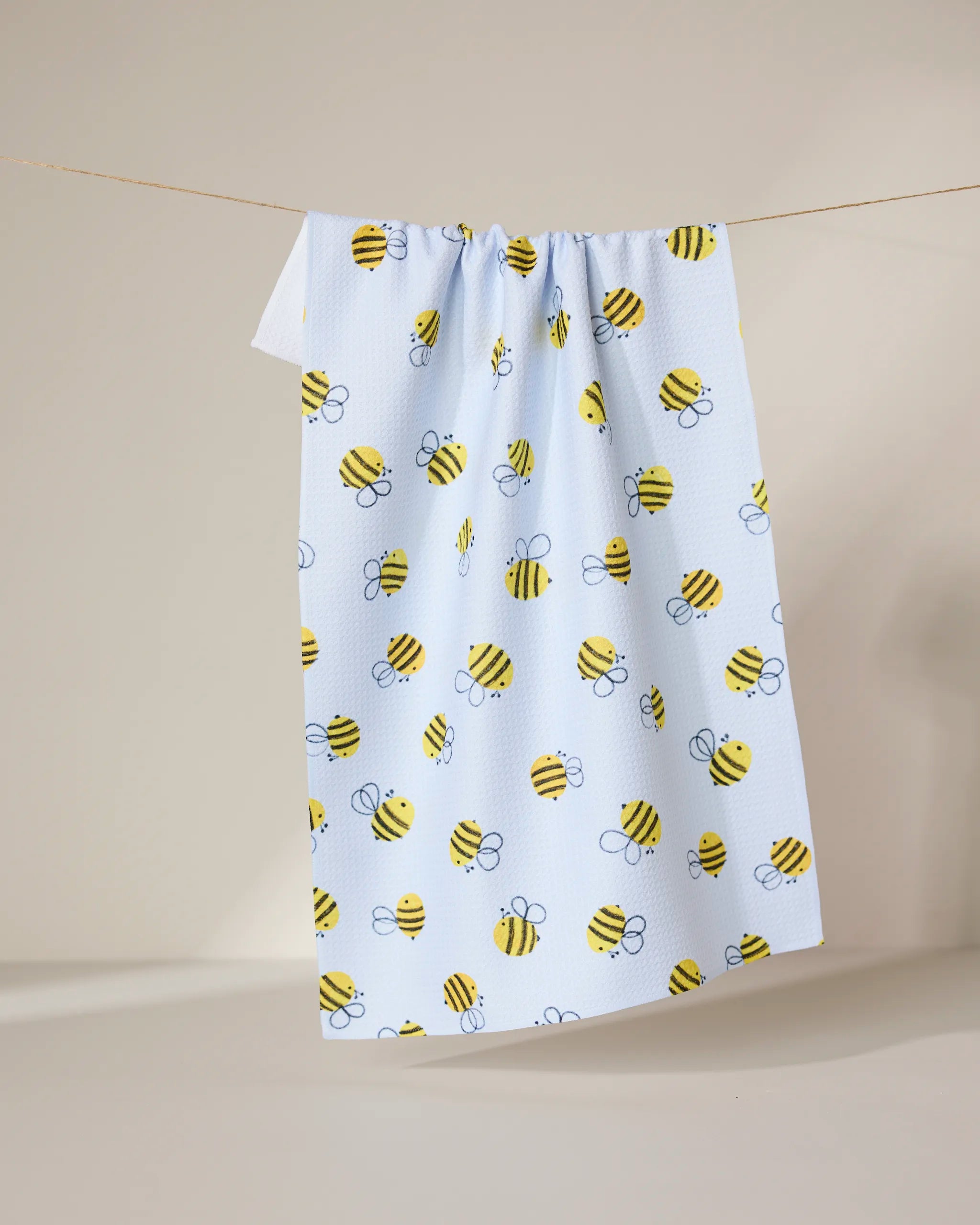 The GEOMETRY Buzzing tea towel, in light blue with a playful yellow and black bumblebee pattern, hangs on a string against a plain, light background.