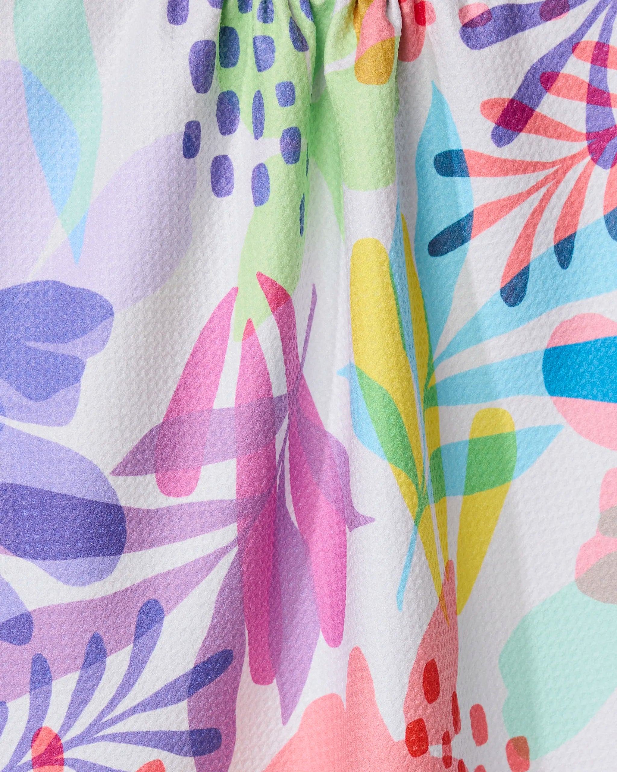 Close-up of GEOMETRYs Botanical Burst kitchen towel, showcasing colorful abstract floral patterns with pink, purple, blue, green, and yellow shapes on textured white recycled fabric.