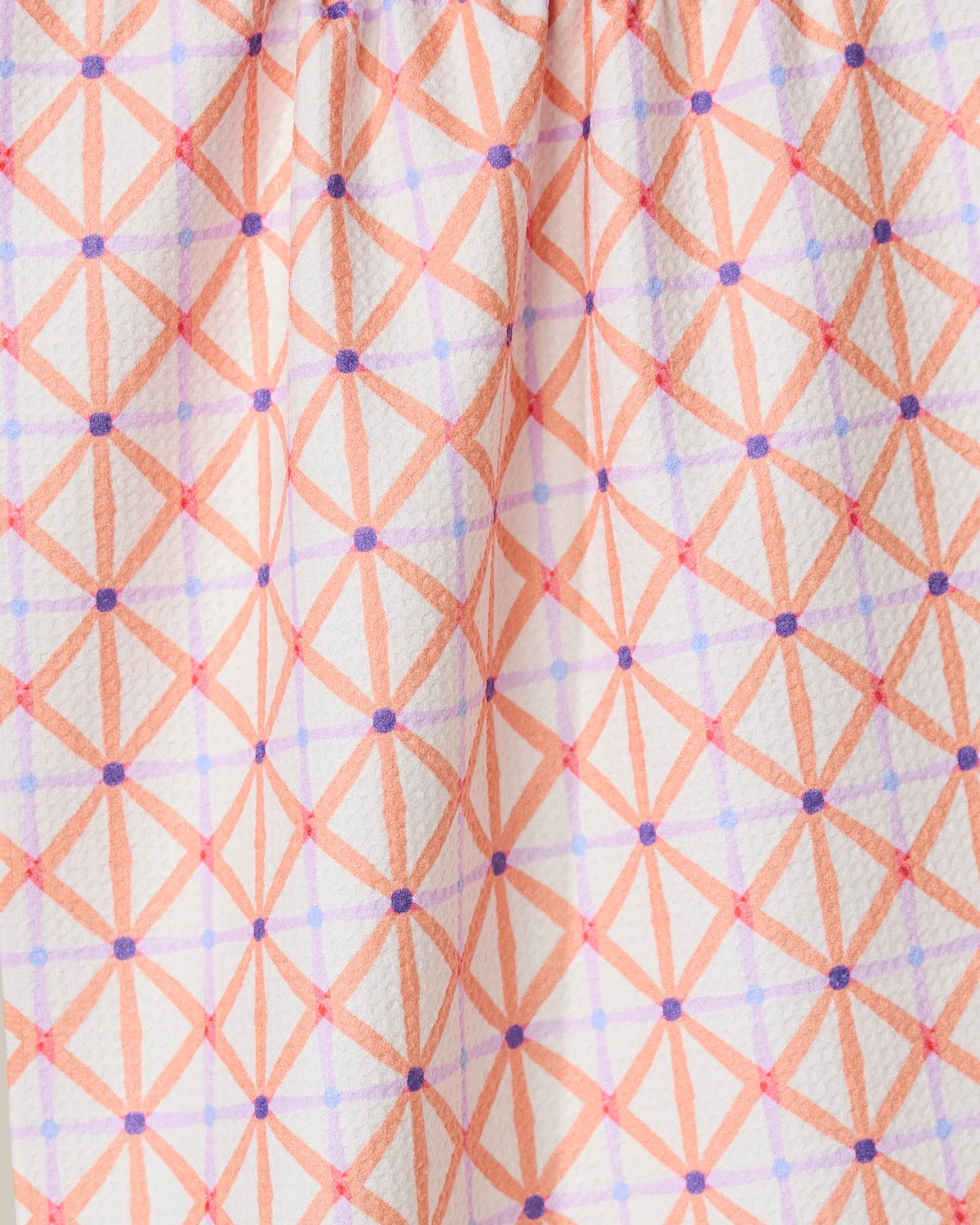 Close-up of GEOMETRYs Anew tea towel made from recycled materials, showcasing a textured white fabric with an orange diamond geometric pattern, accented by intersecting purple lines and dots, gathered at the top.