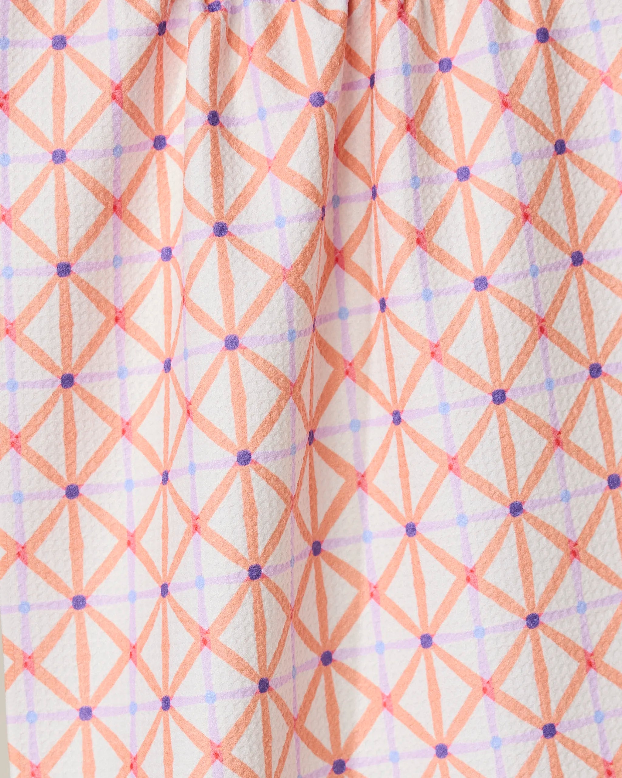 Close-up of GEOMETRYs Anew tea towel made from recycled materials, showcasing a textured white fabric with an orange diamond geometric pattern, accented by intersecting purple lines and dots, gathered at the top.