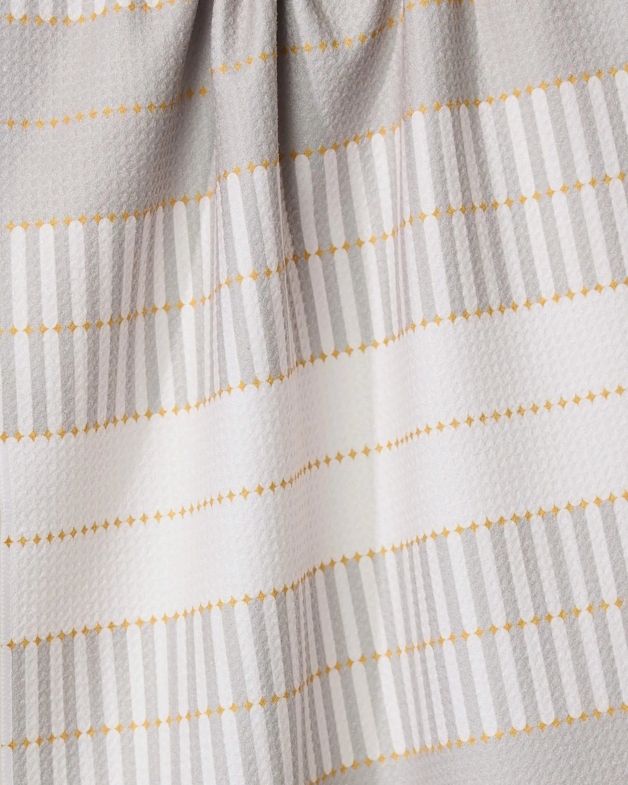 A close-up of the GEOMETRY Bâton Dor absorbent dish towel, featuring horizontal light gray stripes and dotted yellow lines on a textured white background, with gentle fabric folds at the top.