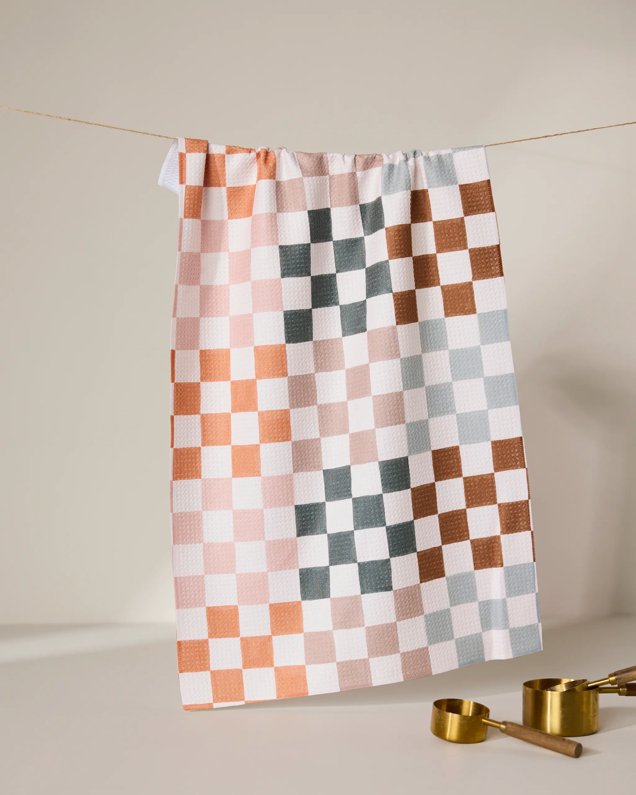 The GEOMETRY Autumn Checkers tea towel, with peach, white, gray, and rust squares, hangs on a string above gold measuring cups. Its ultra-absorbent design stands out against the clean, neutral background.