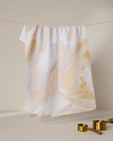The Acete recycled kitchen towel by GEOMETRY, featuring an abstract pastel pattern in beige, white, and light brown, hangs on a thin string with gold measuring cups placed below.