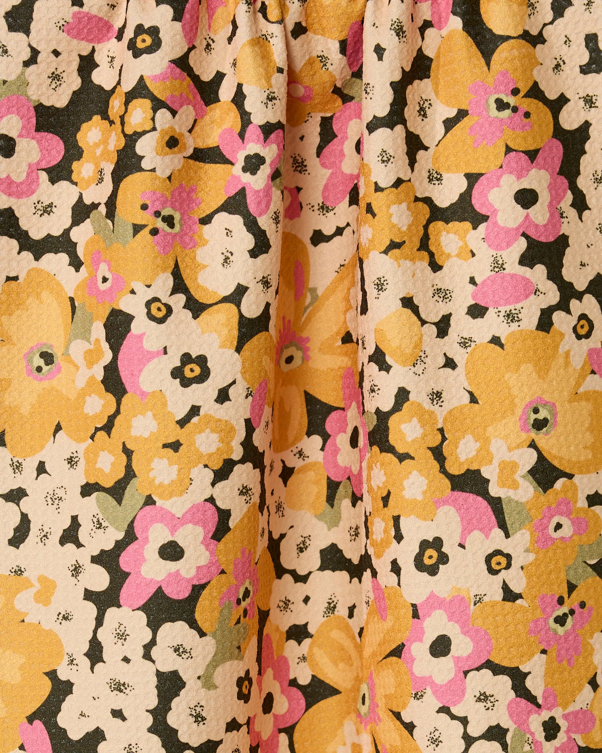 A close-up of the GEOMETRY Beloved Bunch kitchen towel, an eco-friendly option with a vibrant floral pattern in yellow, pink, white, and black on a light background. The fabric features a textured surface and gentle gathered folds.