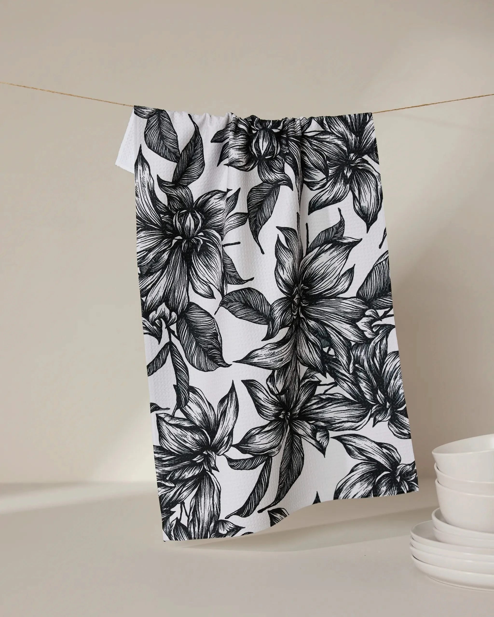 The GEOMETRY Caring tea towel, featuring a bold black floral pattern on ultra-absorbent white fabric, hangs from a string beside stacked white bowls and plates on a neutral background.