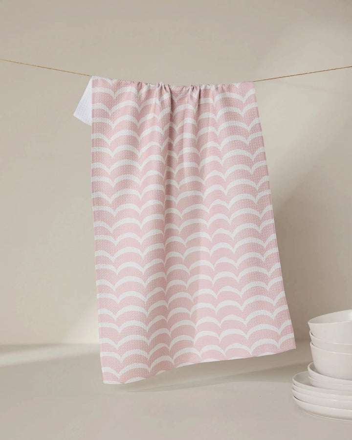 The GEOMETRY Cascading Pink eco-friendly kitchen towel with a scallop pattern hangs on a clothesline, while white ceramic bowls and saucers are stacked nearby on a beige surface against a neutral background.