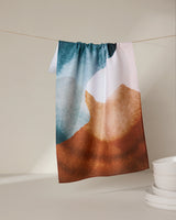 The GEOMETRY Centered kitchen towel features an abstract, watercolor-style design in blue, white, and brown. Made from recycled materials, it adds artistic charm to your kitchen while hanging above stacked white bowls.