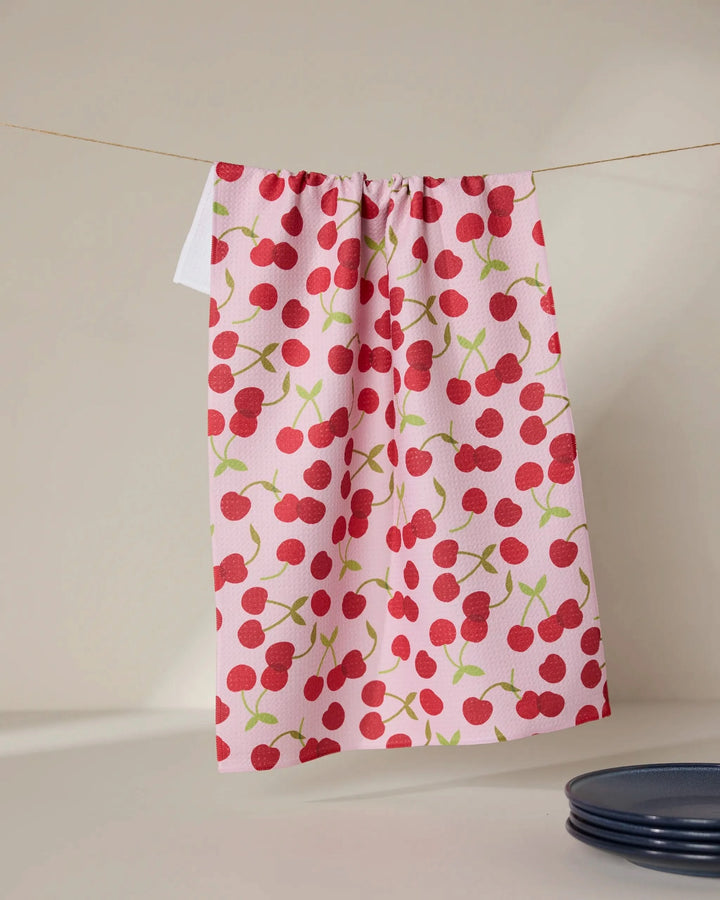 The Cheery Cherries towel by GEOMETRY, featuring a pink ultra-absorbent fabric with a bright red cherry pattern, hangs on a line against a neutral background, while a dark blue plate appears at the bottom right.