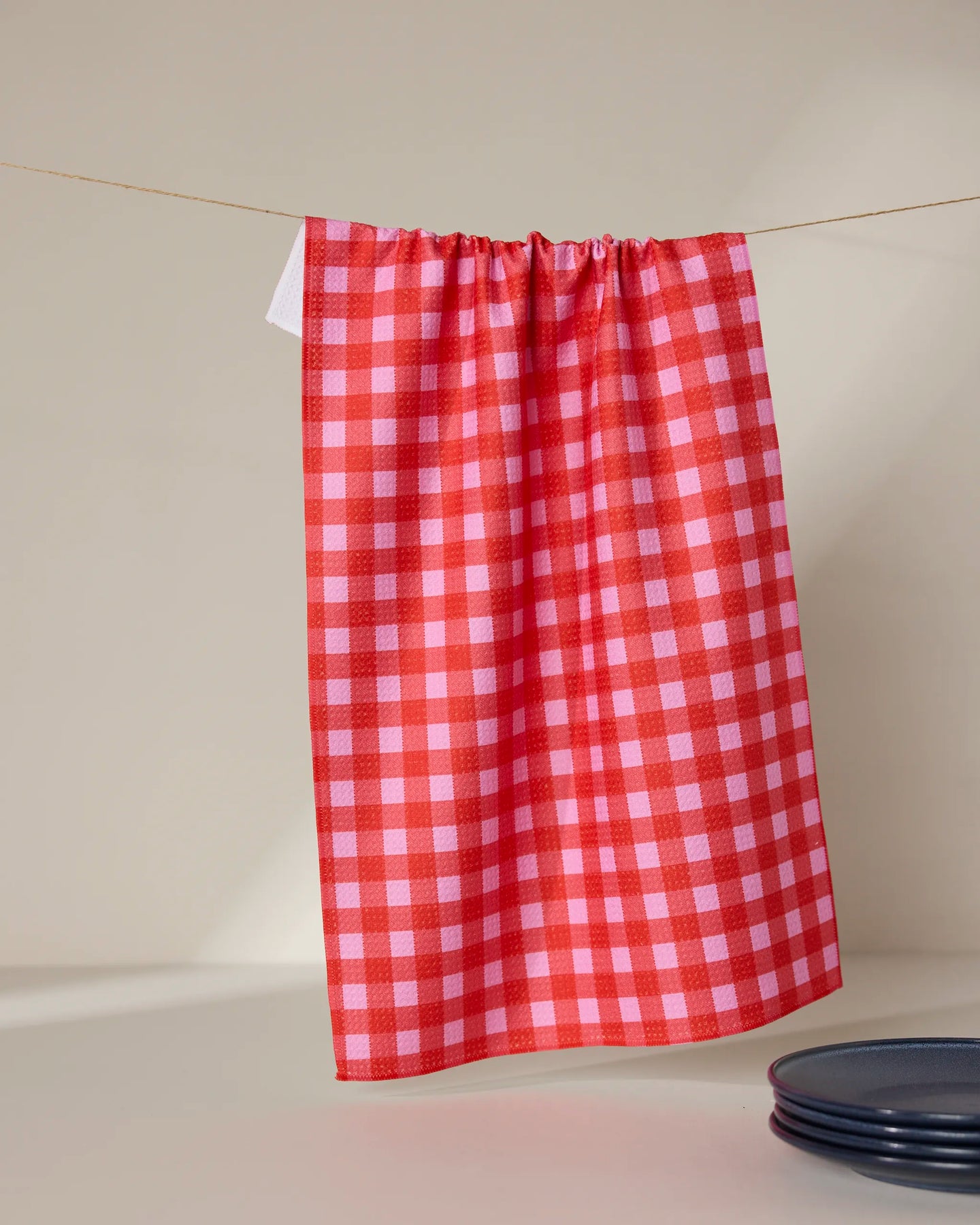 The GEOMETRY Cherry Gingham tea towel hangs on a line, with a stack of dark plates partly visible in the lower right corner, set against a neutral background.