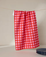 The GEOMETRY Cherry Gingham tea towel hangs on a line, with a stack of dark plates partly visible in the lower right corner, set against a neutral background.