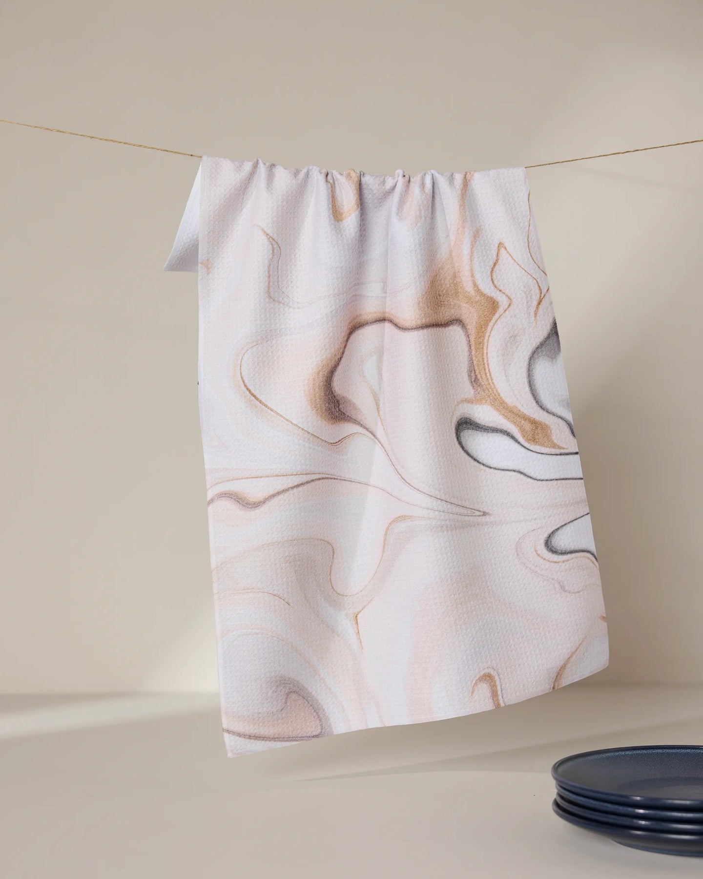 The GEOMETRY Choco tea towel, featuring a light brown and gray marbled pattern, hangs on a clothesline against a beige backdrop with a dark blue plate partly visible at the bottom right.