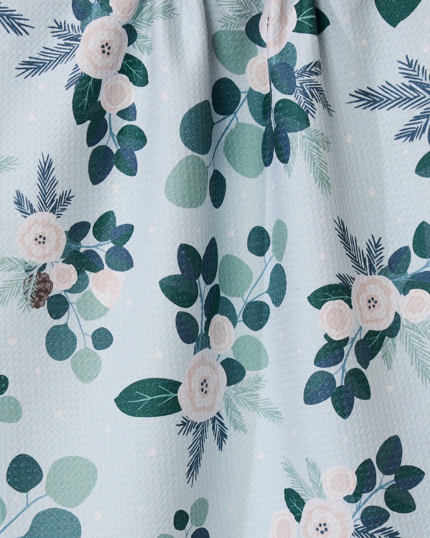 The GEOMETRY Country Winter tea towel is quick-drying and features a charming pattern of white flowers, green leaves, and pine branches on a light blue background with subtle white dots.