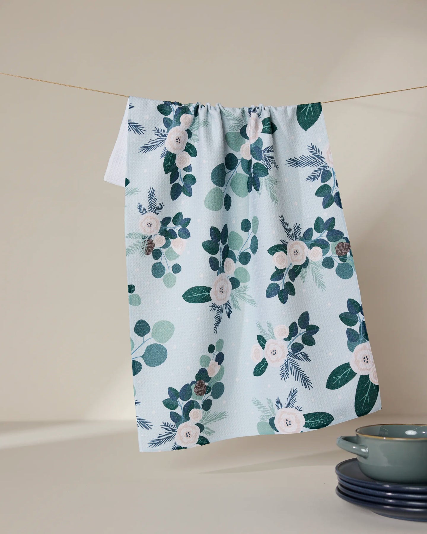 The GEOMETRY Country Winter recycled kitchen towel in light blue, with a floral and leafy green pattern, hangs on a clothesline above neatly stacked blue dishes on a beige surface.