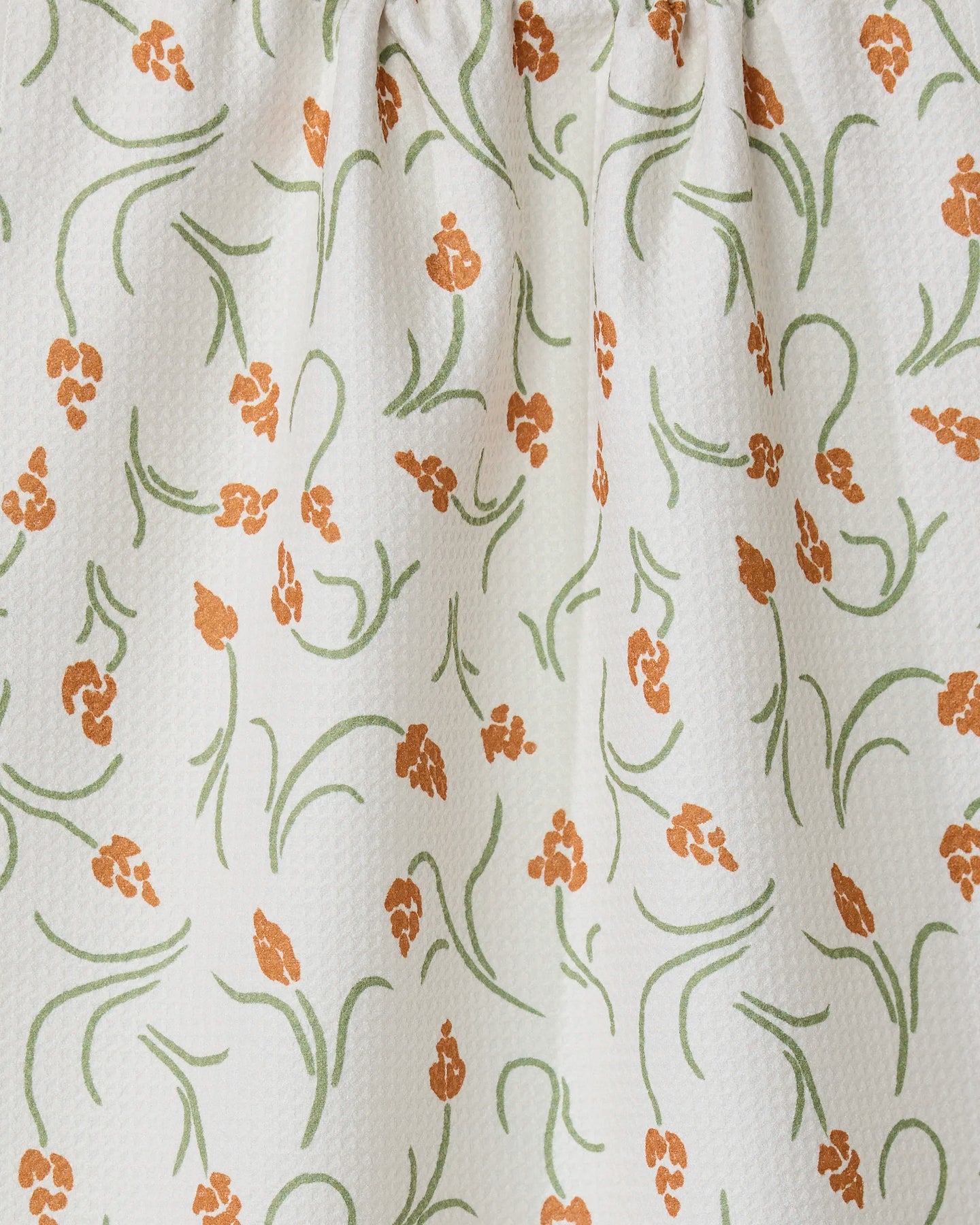The GEOMETRY Crayon Floral tea towel features white textured fabric with orange floral clusters and green curving stems and leaves. Slightly gathered at the top, it offers style, function, and a light, airy look.