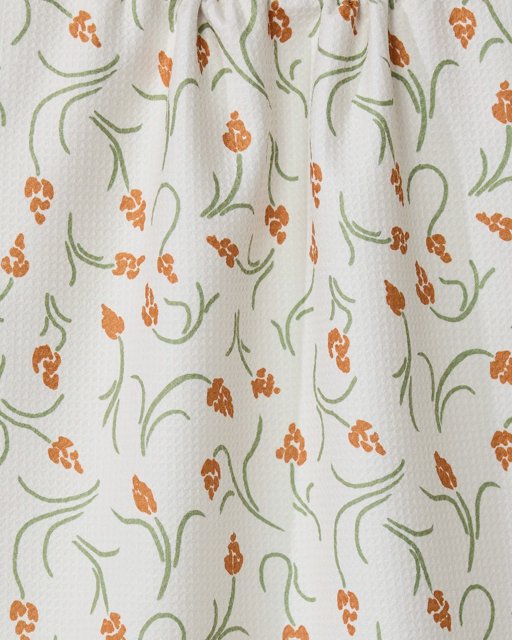 The GEOMETRY Crayon Floral tea towel features white textured fabric with orange floral clusters and green curving stems and leaves. Slightly gathered at the top, it offers style, function, and a light, airy look.