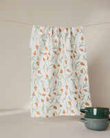 A GEOMETRY Crayon Floral recycled white kitchen towel with an orange and green floral pattern hangs on a line. Two green ceramic bowls are stacked in the bottom right corner against a beige background.