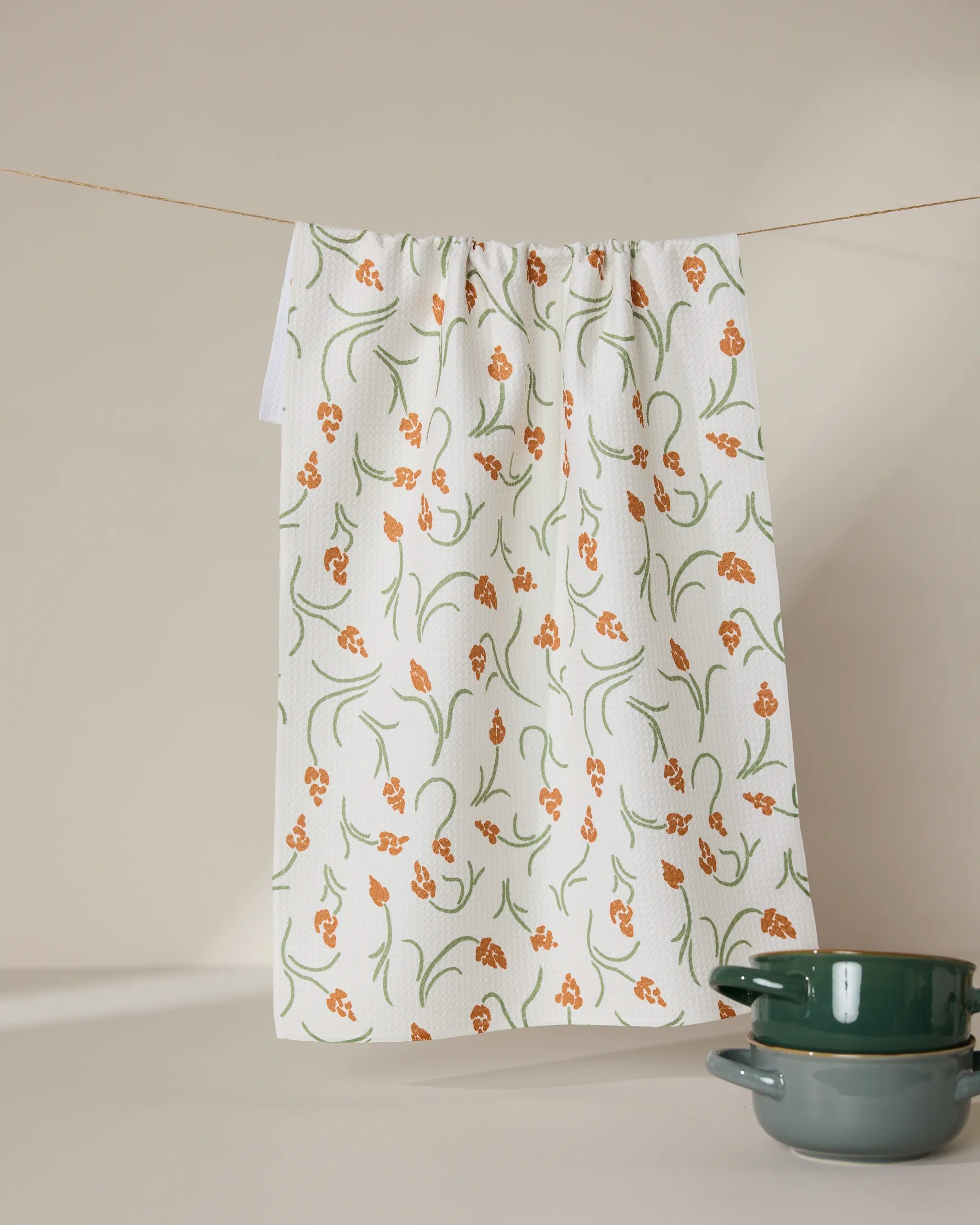 A GEOMETRY Crayon Floral recycled white kitchen towel with an orange and green floral pattern hangs on a line. Two green ceramic bowls are stacked in the bottom right corner against a beige background.