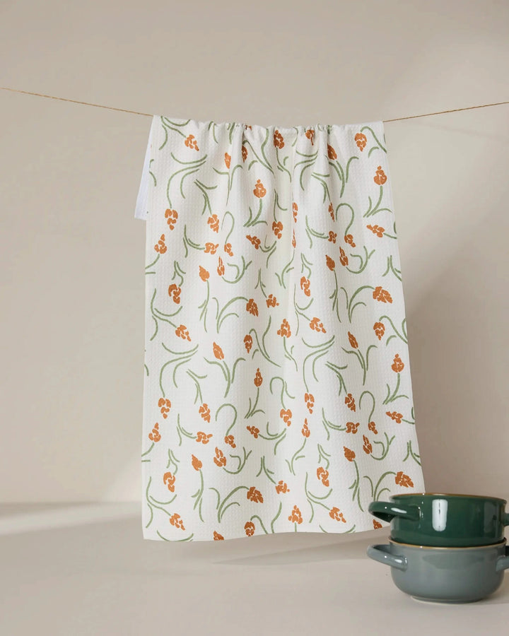 A GEOMETRY Crayon Floral recycled white kitchen towel with an orange and green floral pattern hangs on a line. Two green ceramic bowls are stacked in the bottom right corner against a beige background.