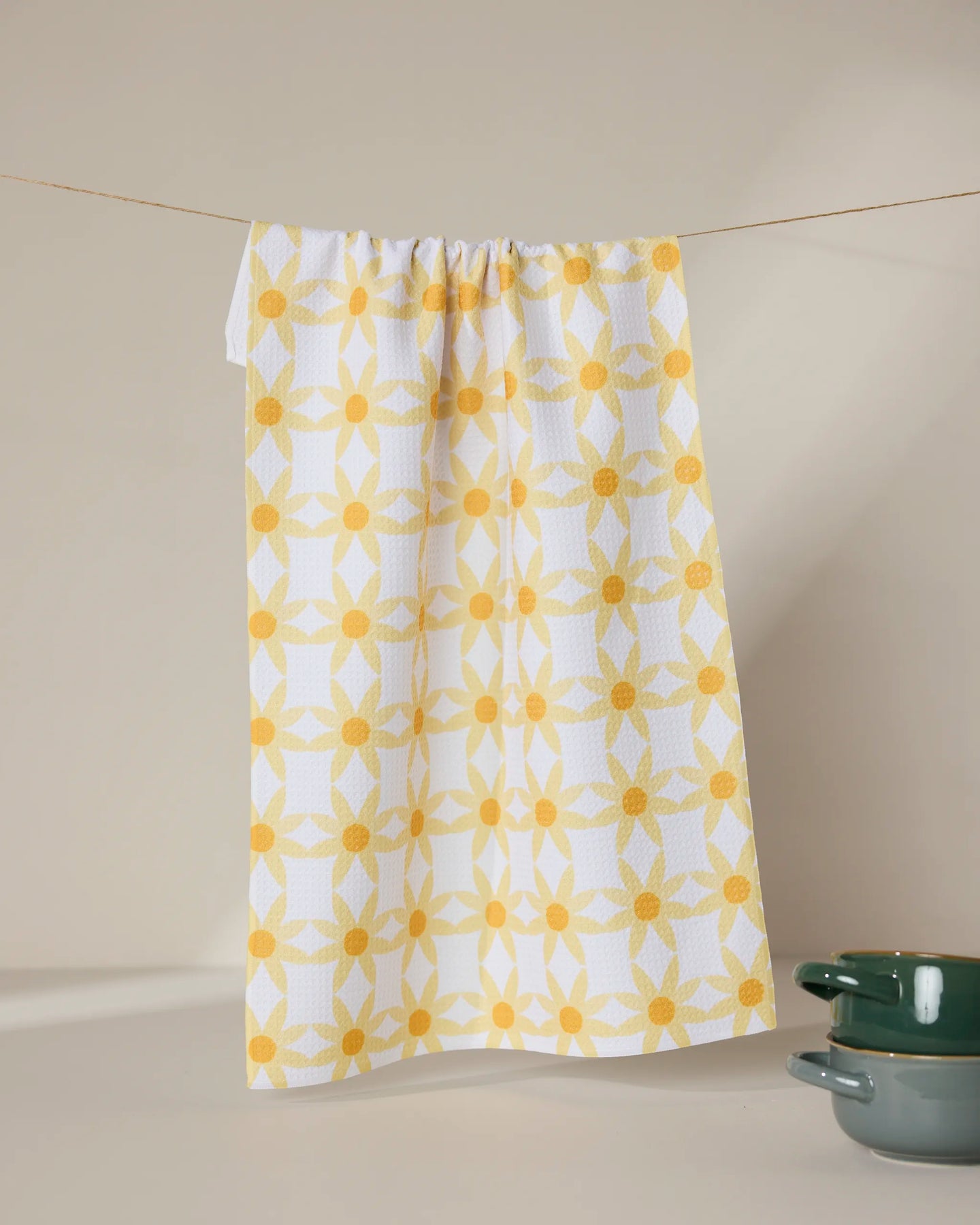 The GEOMETRY Daisies tea towel, featuring a yellow geometric floral pattern and made from recycled materials, hangs on a string. Two green ceramic bowls are stacked in the bottom right corner against a neutral background.