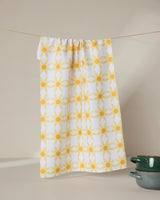 The GEOMETRY Daisies tea towel, featuring a yellow geometric floral pattern and made from recycled materials, hangs on a string. Two green ceramic bowls are stacked in the bottom right corner against a neutral background.