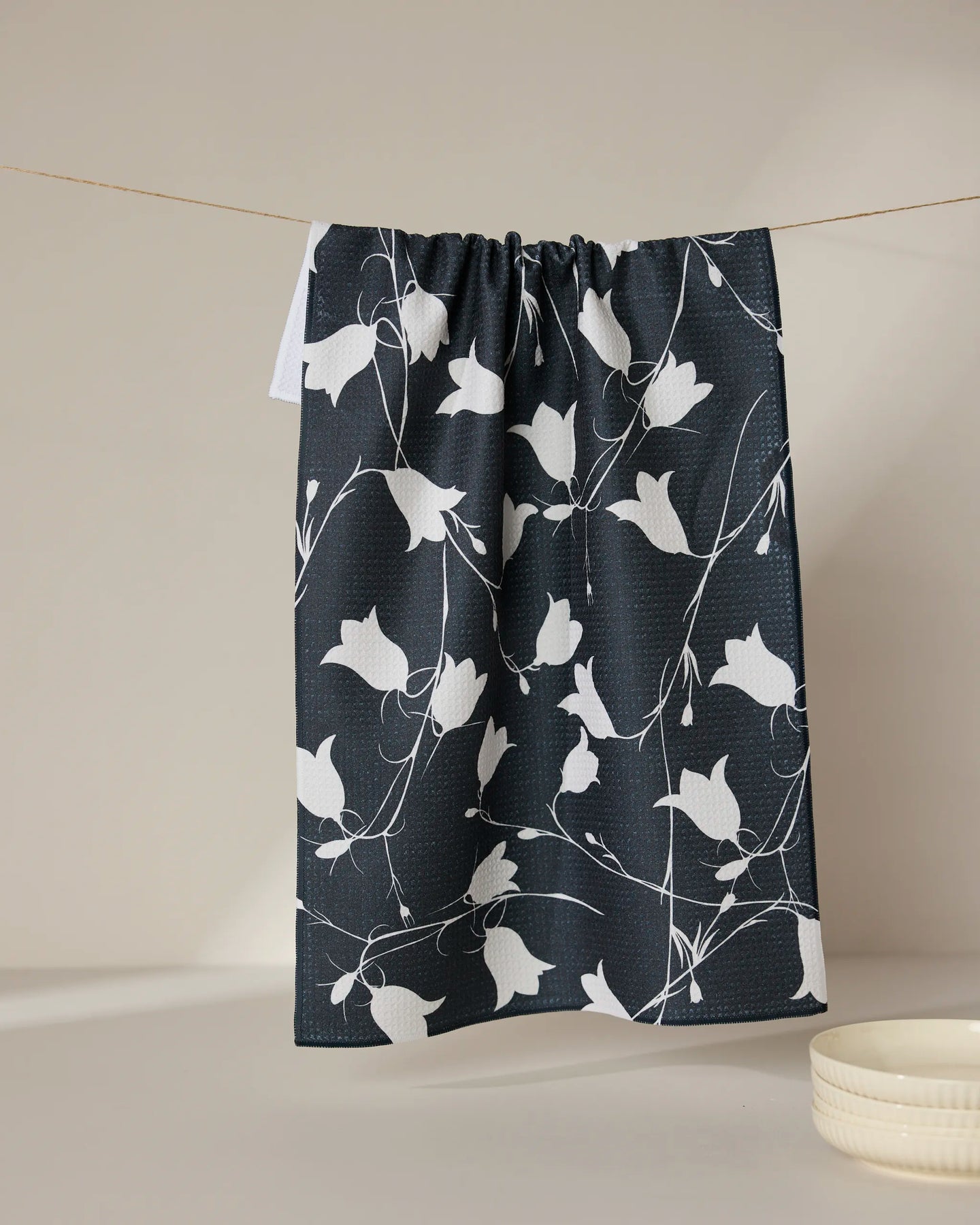 The GEOMETRY Dancing Dark Flora ultra-absorbent towel, featuring a white floral pattern, hangs on a thin line against a neutral background with a small white bowl nearby.