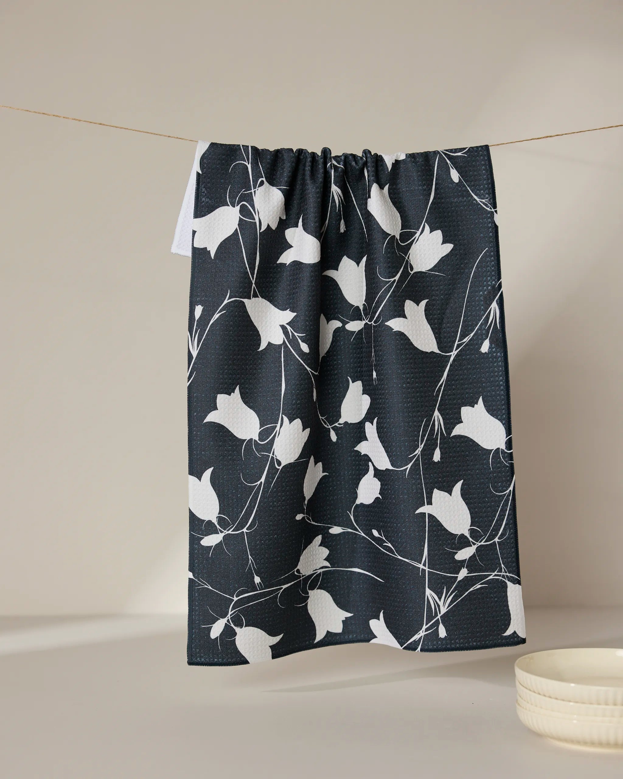 The GEOMETRY Dancing Dark Flora ultra-absorbent towel, featuring a white floral pattern, hangs on a thin line against a neutral background with a small white bowl nearby.