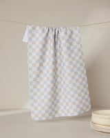 The GEOMETRY Danse Neutre tea towel, featuring a checkered white and light gray design, hangs on a clothesline against a neutral background, with a stack of white plates in the lower right corner.