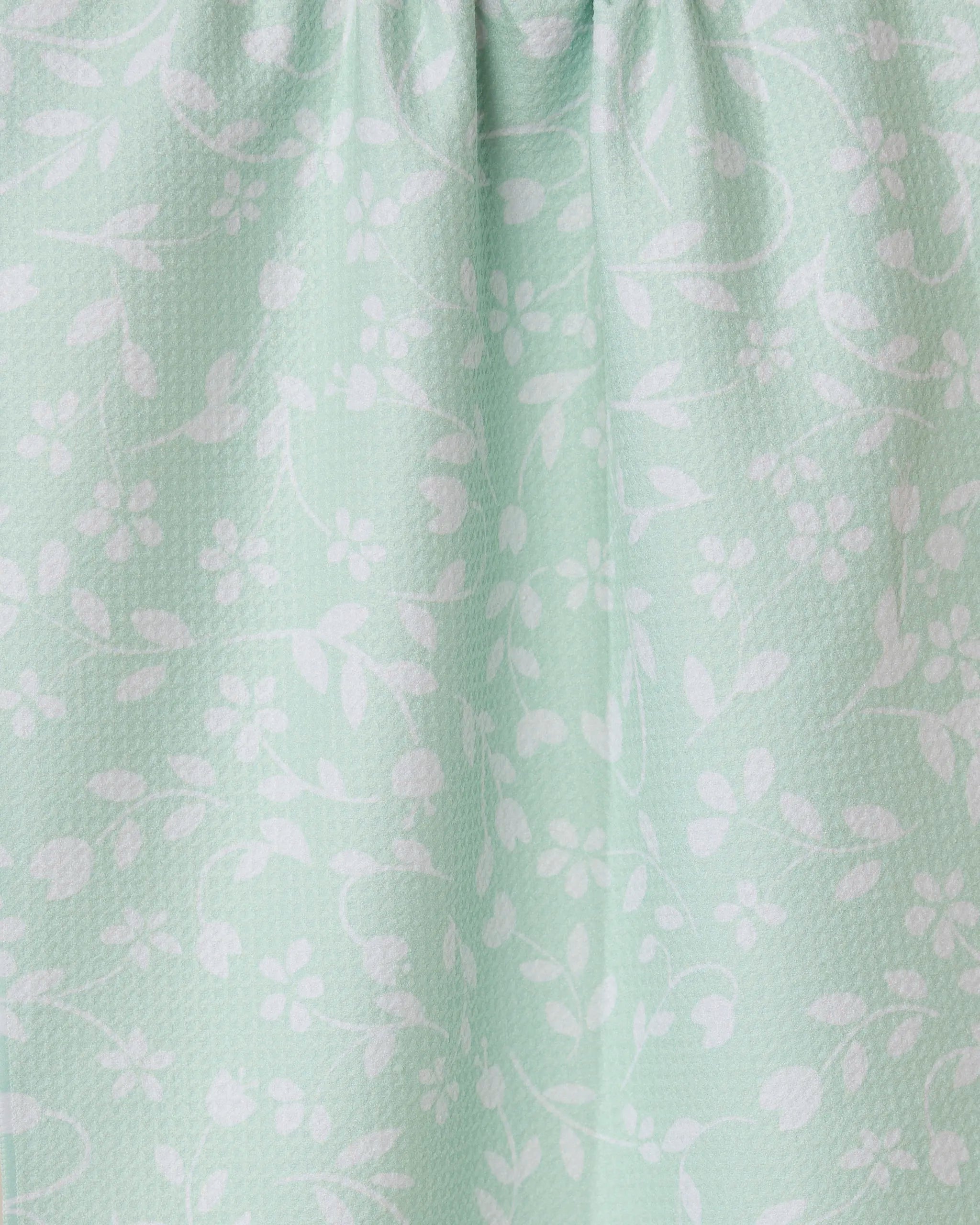 The Dandy kitchen towel by GEOMETRY is light green with white floral and leaf designs. It features a subtle textured pattern and softly gathered fabric at the top for gentle folds.