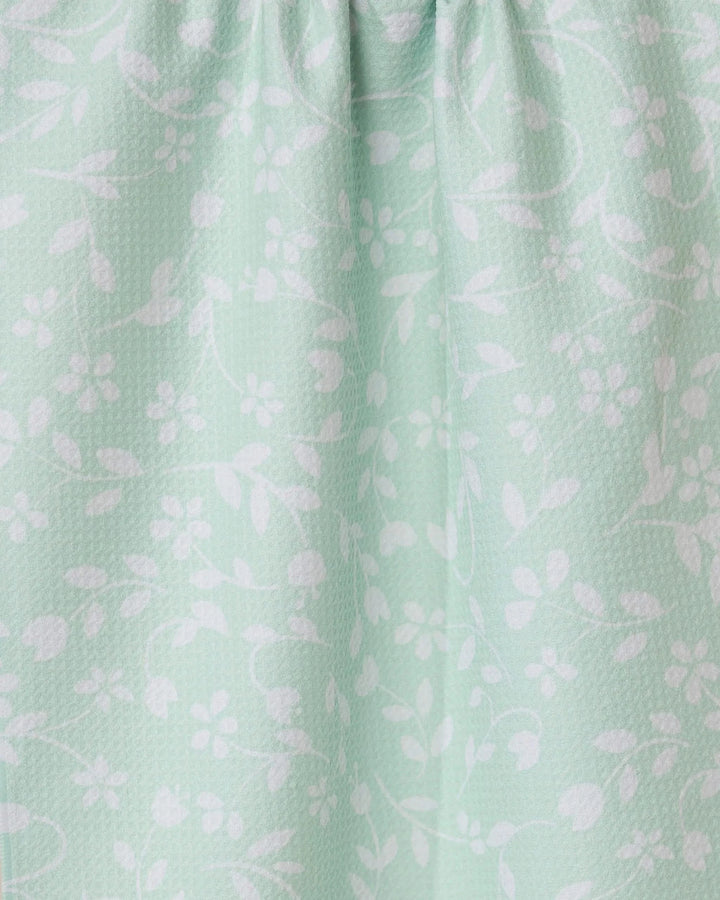 The Dandy kitchen towel by GEOMETRY is light green with white floral and leaf designs. It features a subtle textured pattern and softly gathered fabric at the top for gentle folds.