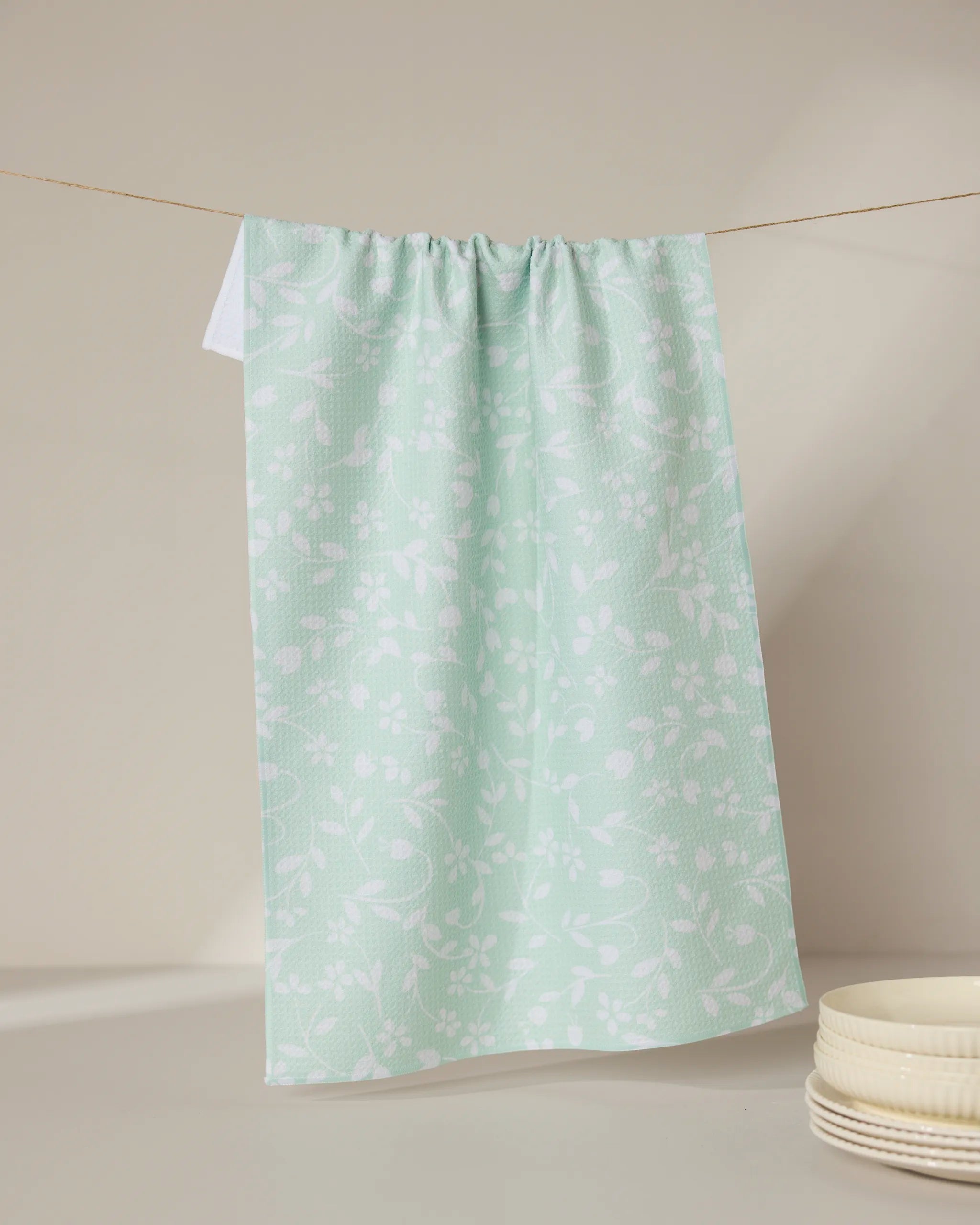 The GEOMETRY Dandy kitchen tea towel, light green with a white floral pattern and made from recycled materials, hangs on a clothesline. Ultra-absorbent and eco-friendly, it’s pictured with white bowls in the lower right corner.