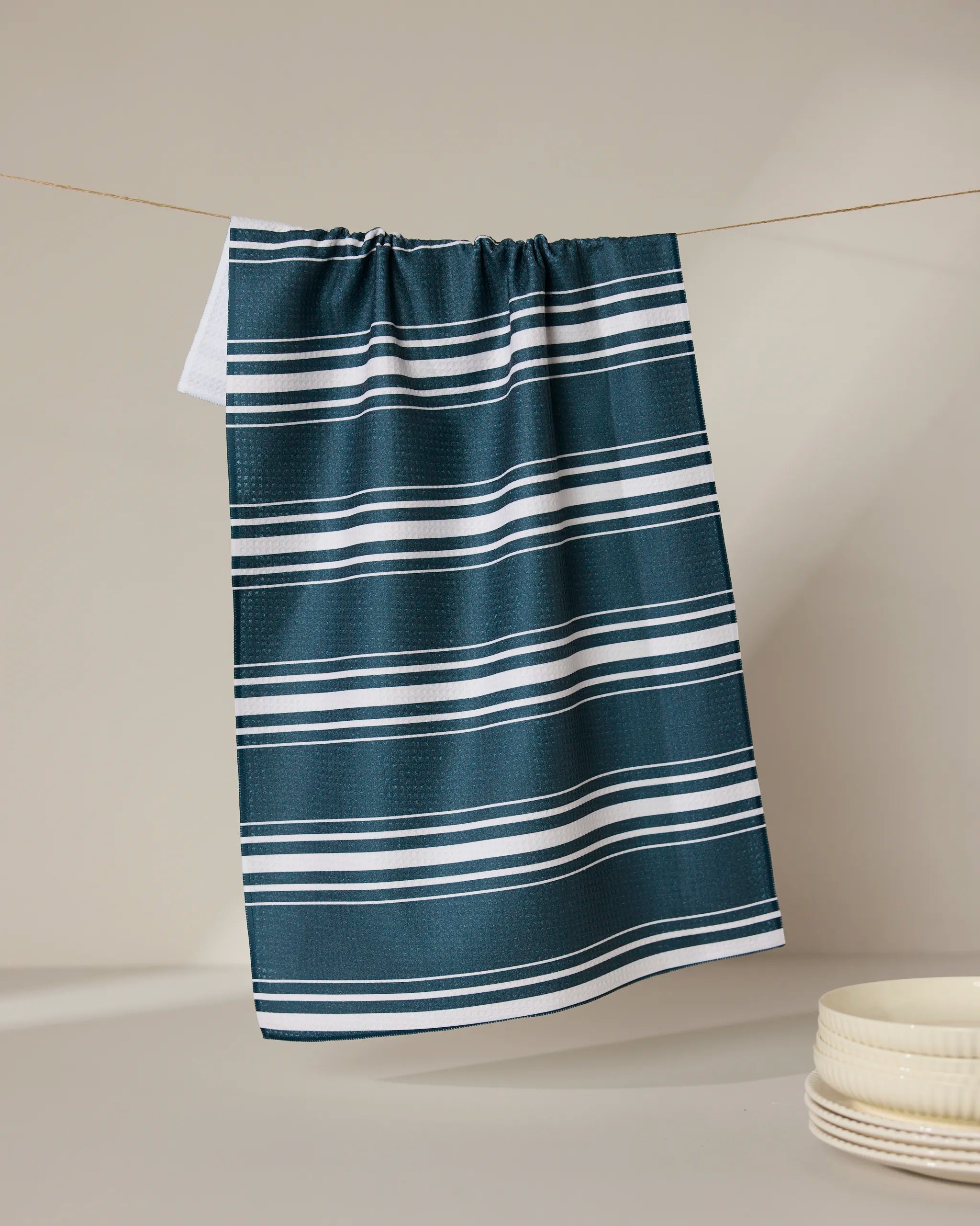 The GEOMETRY Deep Blue Fall recycled kitchen towel, featuring white horizontal stripes, hangs on a string against a neutral backdrop with stacked white ceramic dishes in the lower right corner.