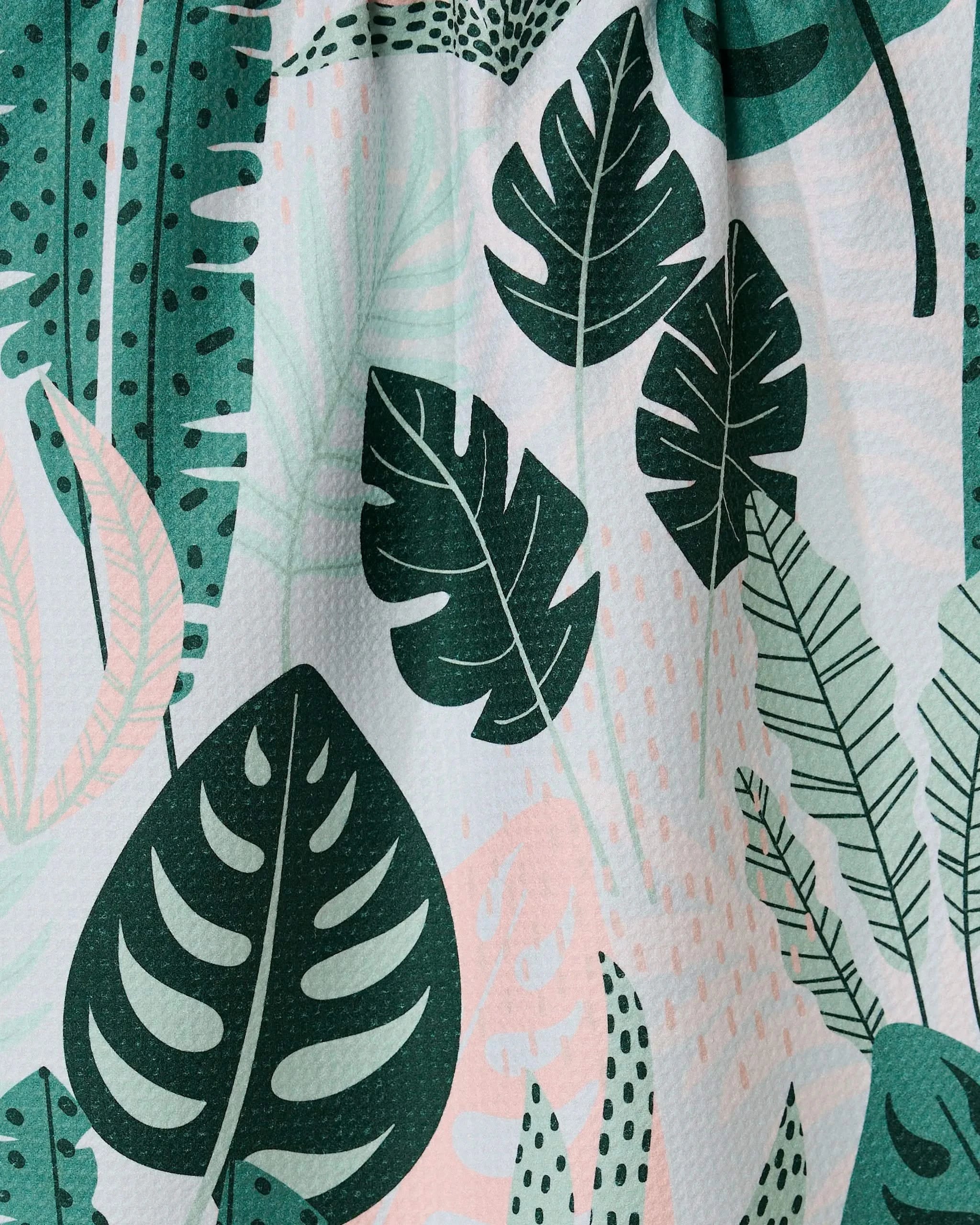 The Diedre kitchen towel by GEOMETRY is an eco-friendly option featuring a variety of green tropical leaf patterns with unique shapes and textures on a light background with soft pink accents.